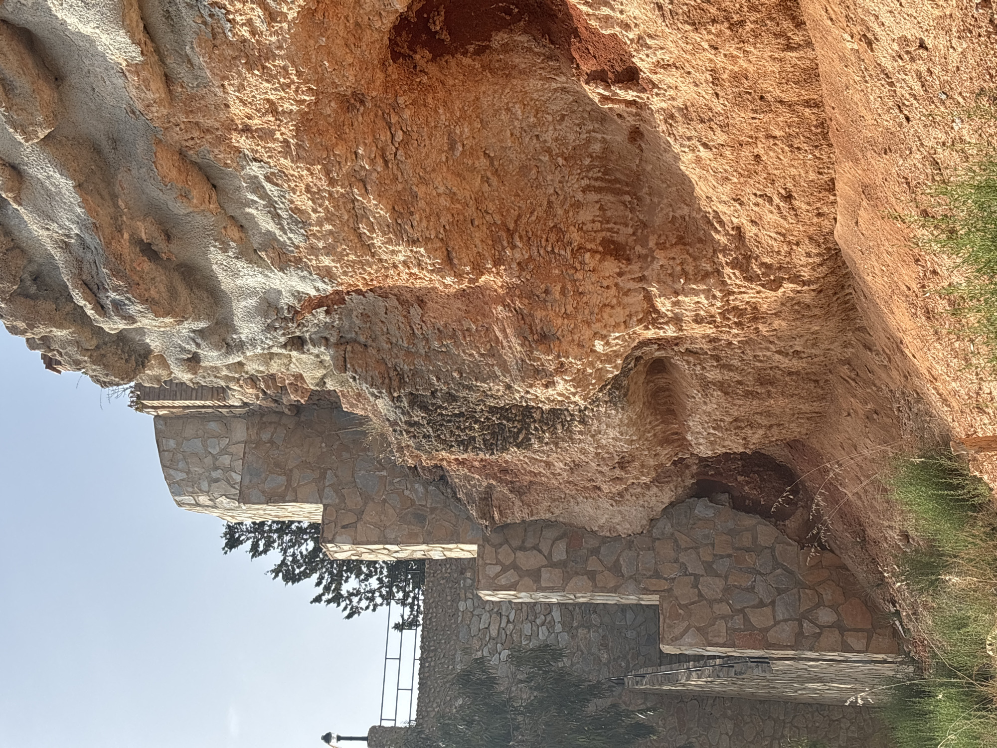 The image captures a section of a cliff face constructed with rough, textured rocks in earthy tones of orange and brown, with patches of lighter grey stone. The rock formation appears weathered and natural, with visible striations and indentations. Below the cliff, there is a series of stone retaining walls and terraces, constructed with irregularly shaped, flat stones. These walls create a tiered structure that ascends the cliff.
Interspersed among the stonework are glimpses of vegetation. On the right side of the frame, dry, wispy grass grows against the rock face. On the left, a dark green tree with needle-like foliage emerges from behind one of the stone terraces. In the background, a clear, pale blue sky suggests fair weather. The lighting indicates it is likely daytime, with sunlight casting shadows and highlighting the textures of the rocks. The perspective is angled, making it difficult to discern specific activities. The scene appears to be a landscape feature, possibly part of a park, garden, or a public space in a naturally rugged environment. There is no visible text in the image.