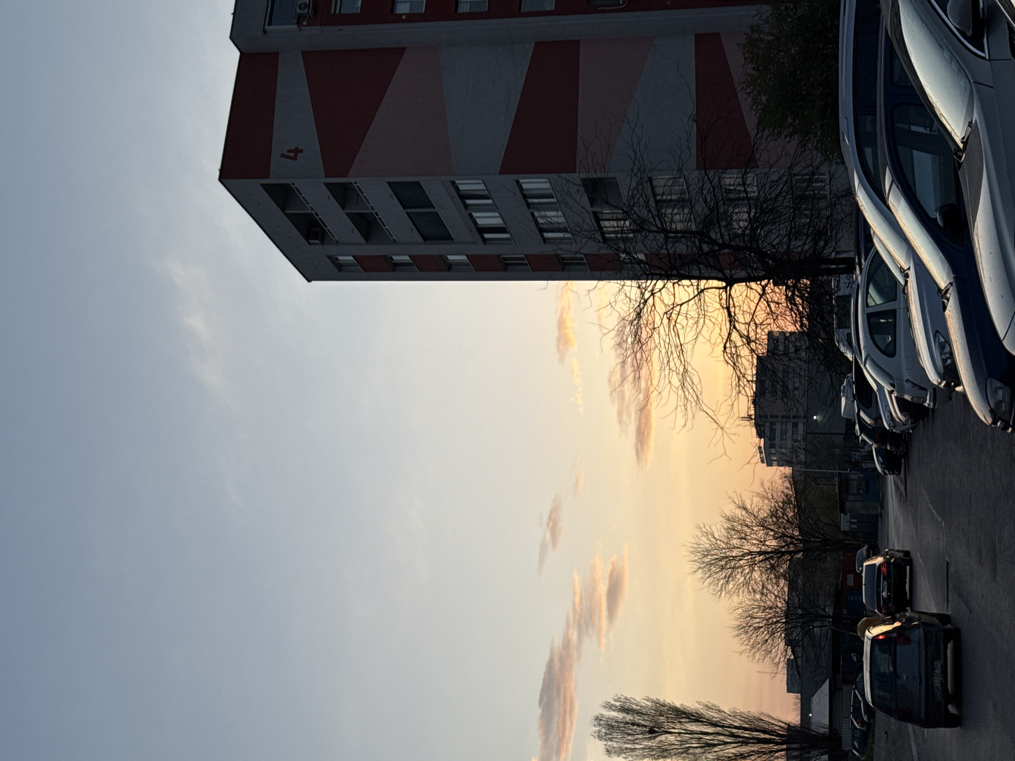 The image depicts a street scene in Sofia, Bulgaria, at dusk. The sky transitions from light blue to orange and yellow near the horizon, with scattered clouds. On the left, a tall, gray building with red and white geometric patterns on its facade rises. It has multiple stories with windows and air conditioning units visible. To the right, a row of parked cars lines the street. A bare tree stands in front of the building and along the roadside, its branches silhouetted against the sky. Further down the street, more buildings and cars are visible. The lighting suggests the photograph was taken during sunset.