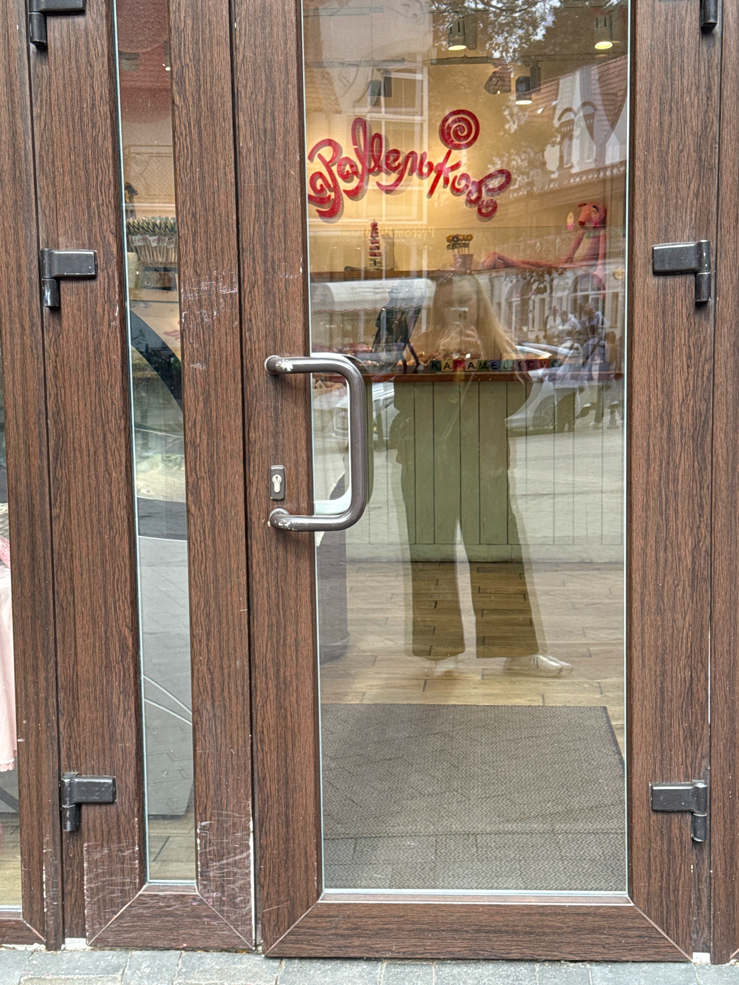 A full shot, eye-level, medium-distance photograph shows the reflection of a young woman taking a selfie in the glass doors of a shop in Svetlogorsk, Russia. The doors are made of dark brown wood with metal hinges and a metal door handle. The woman, who is wearing a light-colored top and wide-leg pants, is reflected from the knees up. She is holding her phone in her right hand to take the picture. In the background of the reflection, there is a shop with a sign that says "КАРАМЕЛЬ" in large, red, cursive letters. There are also some stuffed animals and candies visible inside the shop. The overall lighting is bright, with a slight yellow cast.