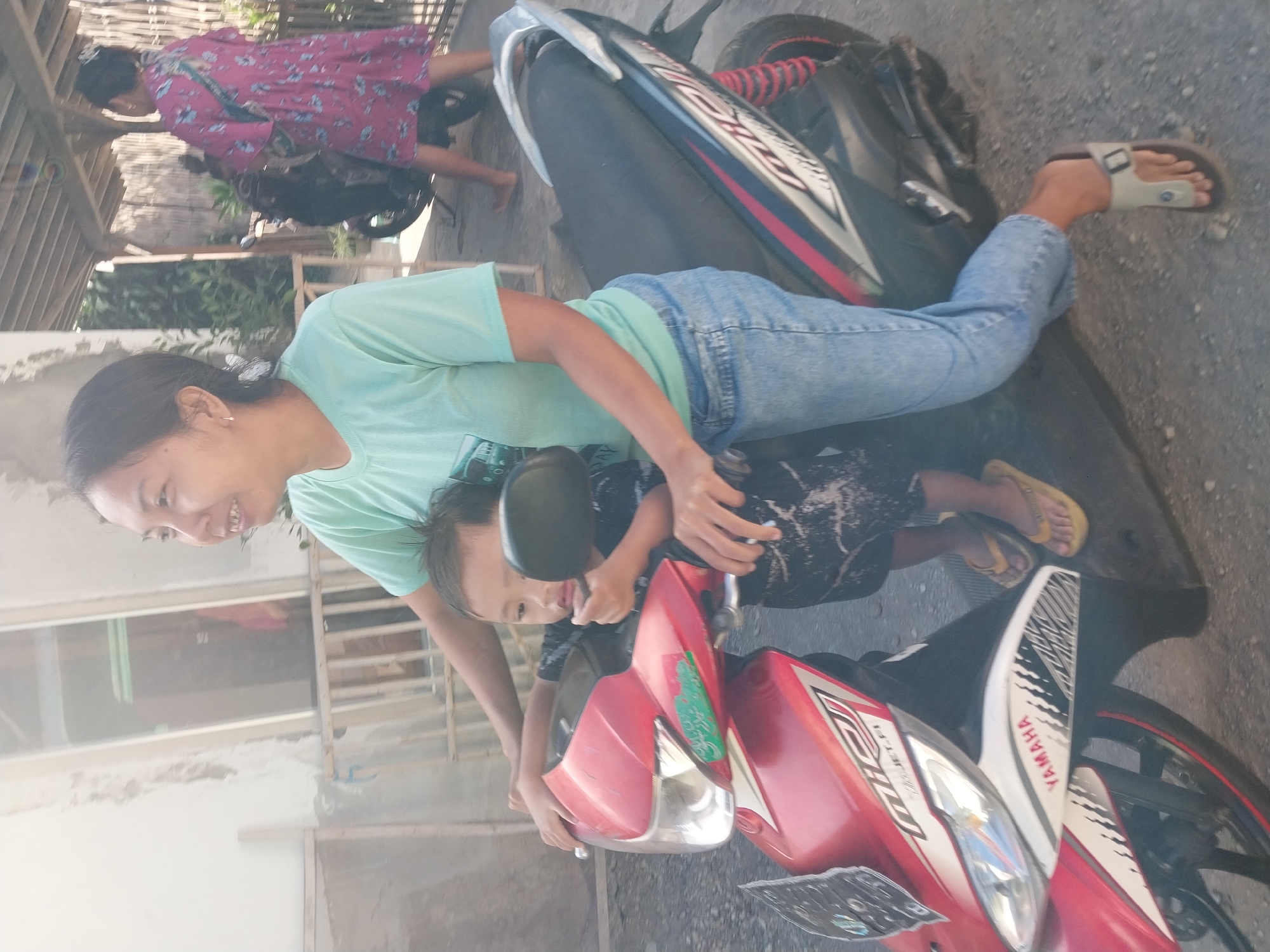 A woman in a light blue t-shirt and blue jeans sits on a red and black scooter. She is holding a young child, who is wearing a black patterned t-shirt and dark shorts. The child is leaning against the woman and holding onto the scooter's handlebars. The woman is looking to her left and smiling. In the background, another woman in a pink floral dress is walking away from the scooter. The scene appears to be outdoors, with a building and a covered structure in the background. The ground is a gravelly or dirt surface. This image was taken in Nglegok, Indonesia.