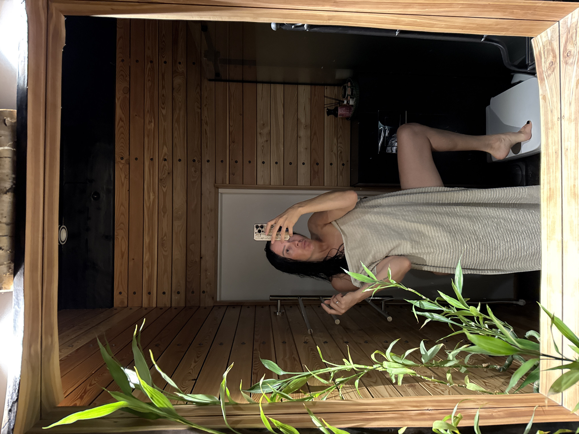 The image shows a reflection in a mirror of a woman taking a selfie in what appears to be a sauna or spa-like room. The room has wooden paneled walls and floor. The woman is partially covered by a light-colored towel and is holding a smartphone to take the picture. Her legs are visible, and she is wearing a ring on her left hand. In the background of the reflection, a toilet and a black wall are visible, along with some shelf items and what looks like a diffuser. There are also some green plants in the foreground of the mirror, which are real and not part of the reflection. The lighting suggests it is daytime, but the interior setting makes it difficult to determine the exact time of day. The overall mood seems relaxed and private. No visible text is present in the image.
