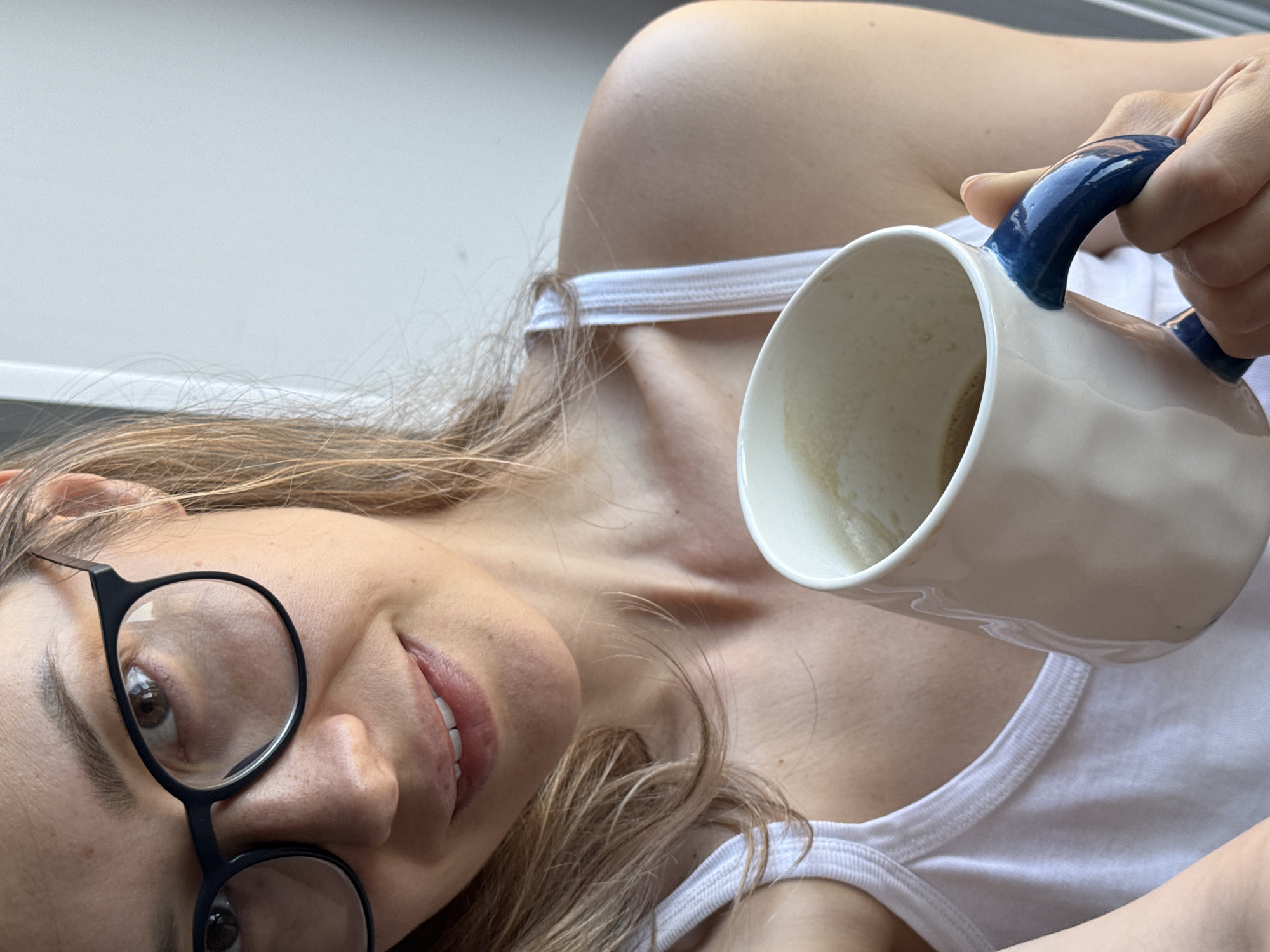 A woman with light brown hair is lying down, wearing a white tank top and black-rimmed glasses. She's holding a white coffee mug with a blue handle in her right hand, nearly empty. The background appears to be a light-colored wall or surface. There is no indication or evidence to suggest that this image was taken in the city of Chaikovsky, Russia.