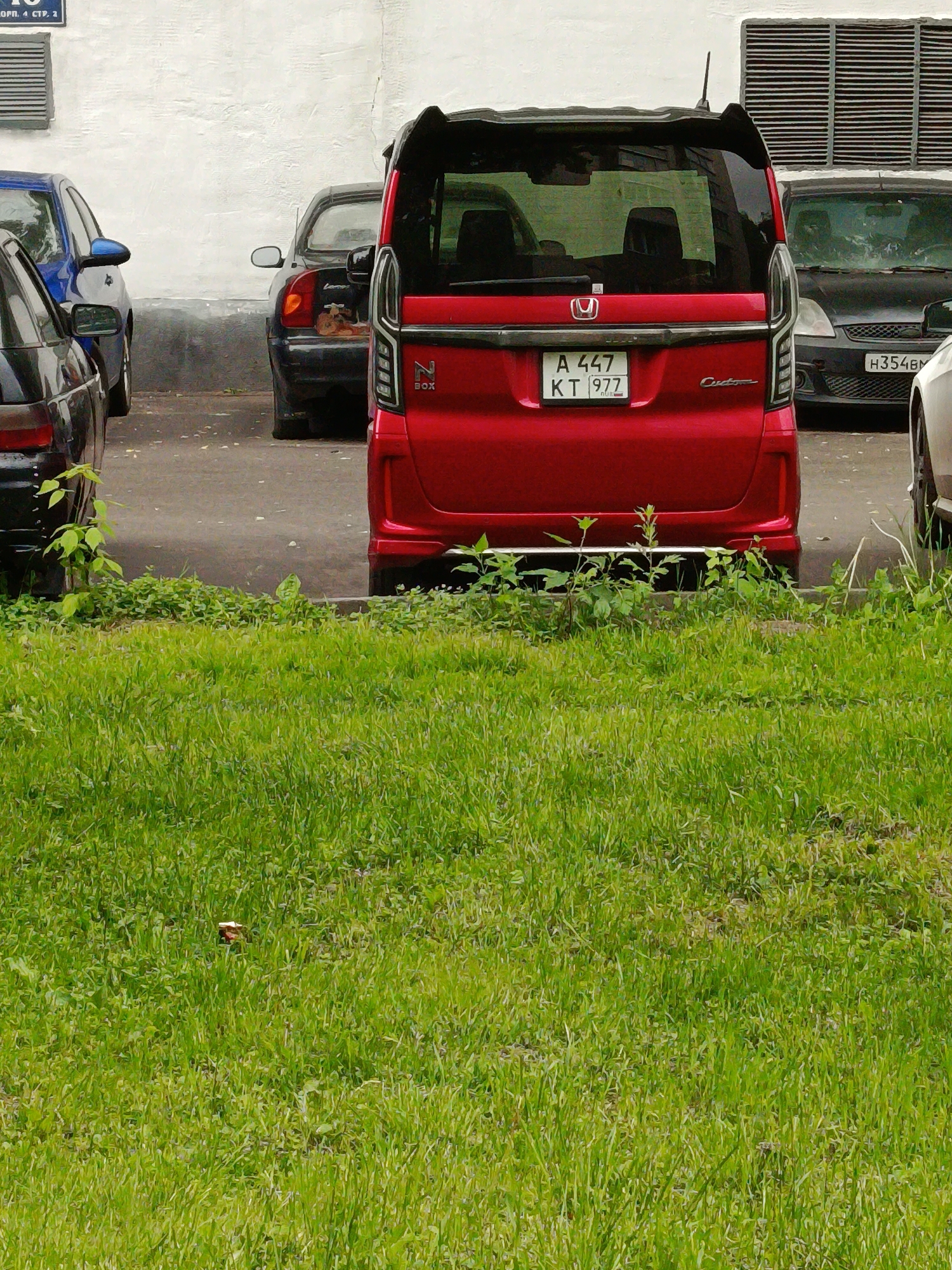 The image captures a scene in an urban parking area, with several cars parked on a paved surface. In the foreground, a vibrant green grassy patch occupies the lower half of the frame.
The central subject is the rear of a red Honda N-Box Custom minivan, parked facing away from the viewer. Its license plate, "A 447 KT 977 RUS," is clearly visible, with "977" indicating a Moscow registration region. The minivan also features "N BOX" and "Custom" badging, along with the Honda emblem. It has a rear spoiler and vertical taillight clusters.
To the left, a black sedan is partially visible, followed by the rear quarter of a blue sedan. To the right of the red minivan, a dark-colored car, possibly a hatchback, is parked, with a partially visible Russian license plate, "H354BH". The front corner of a white vehicle is also seen on the far right edge of the frame.
The background consists of a plain white building wall. On the upper right, a large industrial-style vent or grille is mounted on the wall. A partial sign on the building's upper left section reads "ОБР. 4 СТР. 1", likely part of an address or building designation.
The scene depicts a daytime setting, with no direct sunlight visible, suggesting an overcast day. The visible Russian license plates and building text confirm the location as Moscow, Russia.