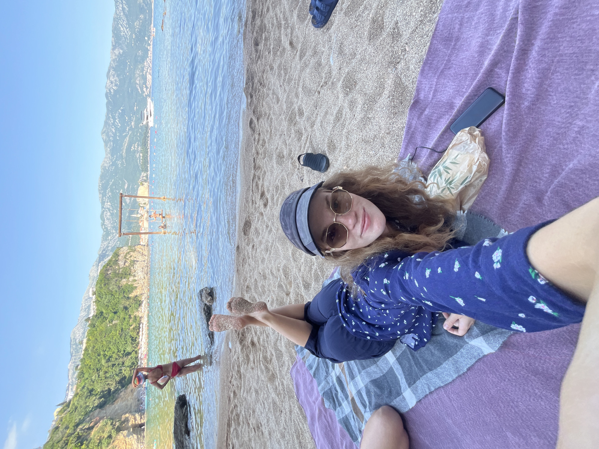 A woman lies on a beach blanket with a phone and paper bag nearby. She wears a blue floral shirt, dark shorts, sunglasses, and a cap. The blanket is purple with a grey and white plaid pattern. In the background, there's a beach with people, the Adriatic Sea, and mountains.
The location is likely a beach near Budva, Montenegro, evidenced by the mountainous coastline and clear water. A person in a red swimsuit is walking in the shallow water, and a swing set is visible in the water further out.
