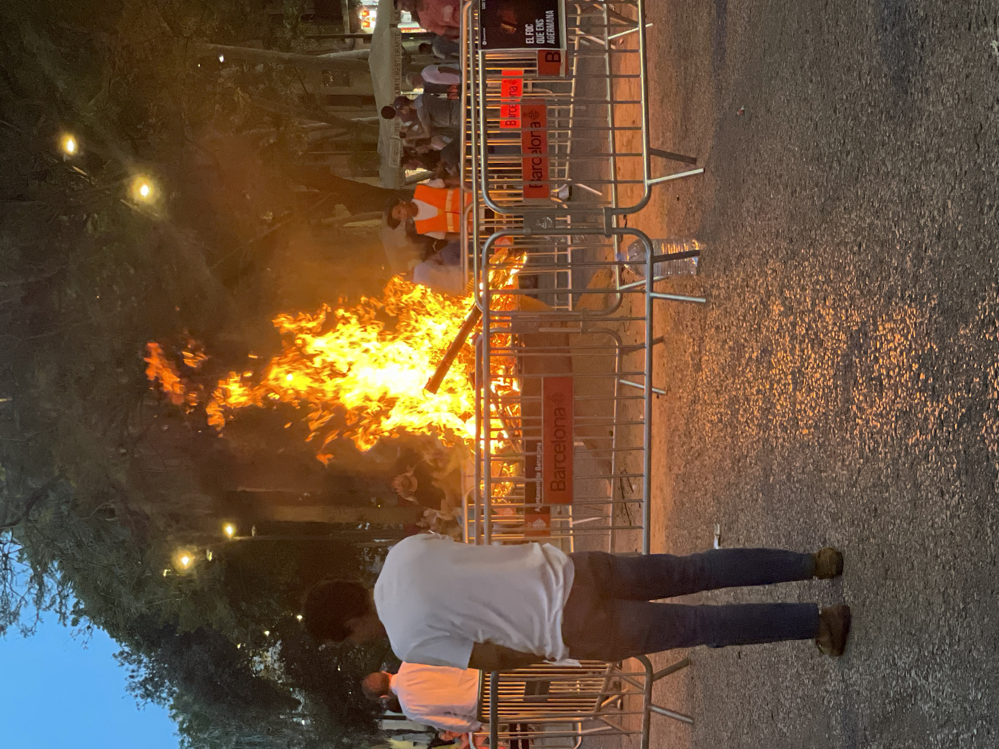 The image depicts an outdoor scene at dusk, possibly a festival or public gathering, centered around a large bonfire. Several people are visible, including one in the foreground standing with their back to the camera, wearing a white t-shirt and jeans. Another person in an orange safety vest is partially visible behind a metal barrier. The bonfire is the dominant element, with bright orange and yellow flames consuming debris.
The scene is cordoned off by metal crowd control barriers. Text visible on the barriers reads "Ajuntament de Barcelona" and "Barcelona," indicating the location is Barcelona, Spain. A sign at the top also reads "EL FOC QUE ENS AGERMANA," which translates to "The fire that unites us." This suggests the event might be related to a local tradition or celebration involving fire.
The setting appears to be a public space, possibly a street or plaza, with trees and building facades visible in the background. Streetlights are also present, contributing to the dusk lighting. The overall atmosphere seems lively, with the presence of a bonfire and a crowd suggesting a celebratory or communal event.