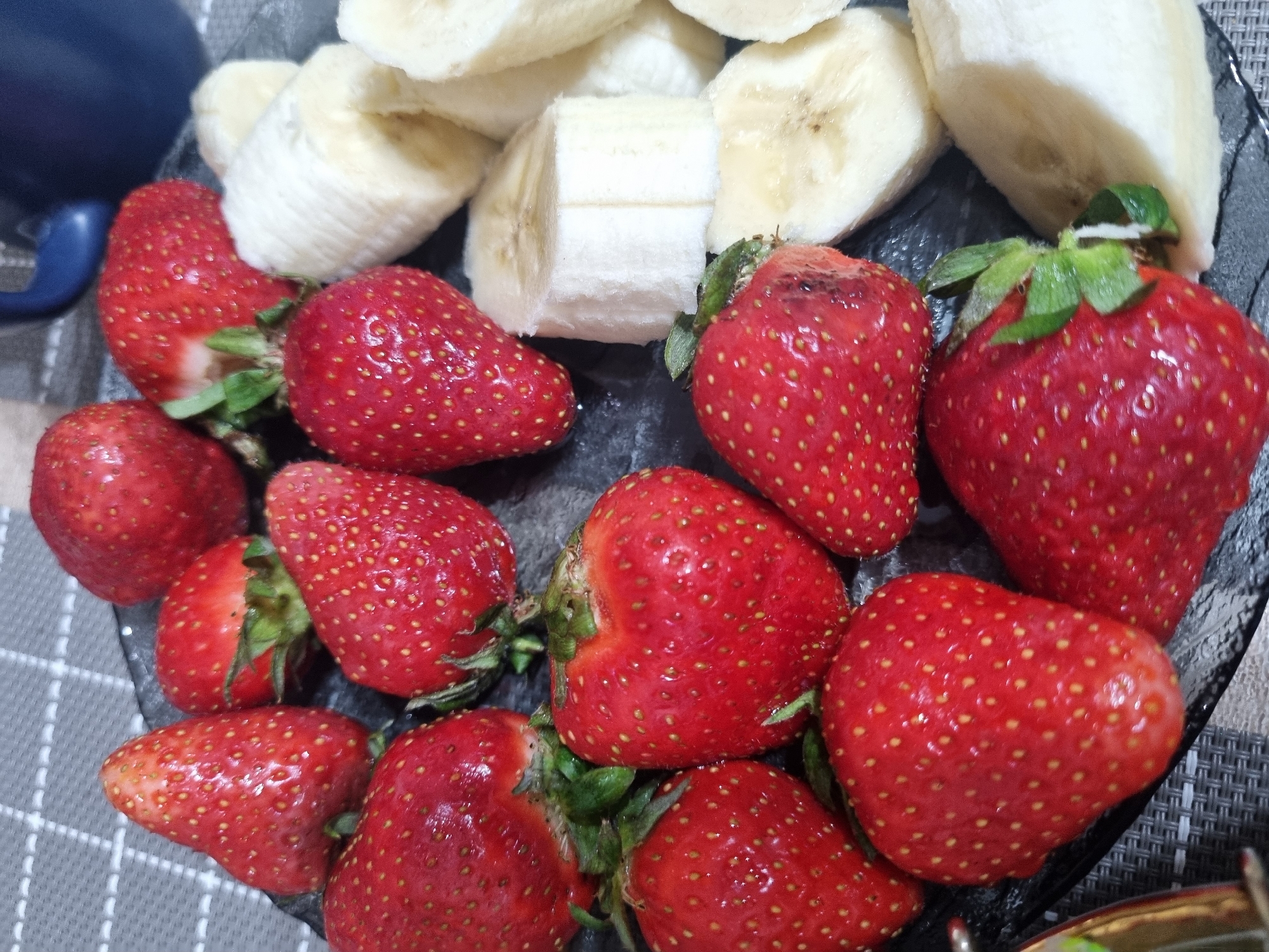 A close-up, overhead shot reveals a plate of strawberries and sliced bananas. The strawberries are bright red and arranged haphazardly, their green stems visible. The bananas are sliced into thick rounds and piled to the side of the strawberries. The plate is a dark, possibly black, color. A blue object, possibly a mug or bowl, is partially visible in the upper left corner. The plate sits on a surface with a grey and white patterned design. The location is unknown, and there is no indication that the image was taken in Sochi, Russia. There are no people visible in the image.