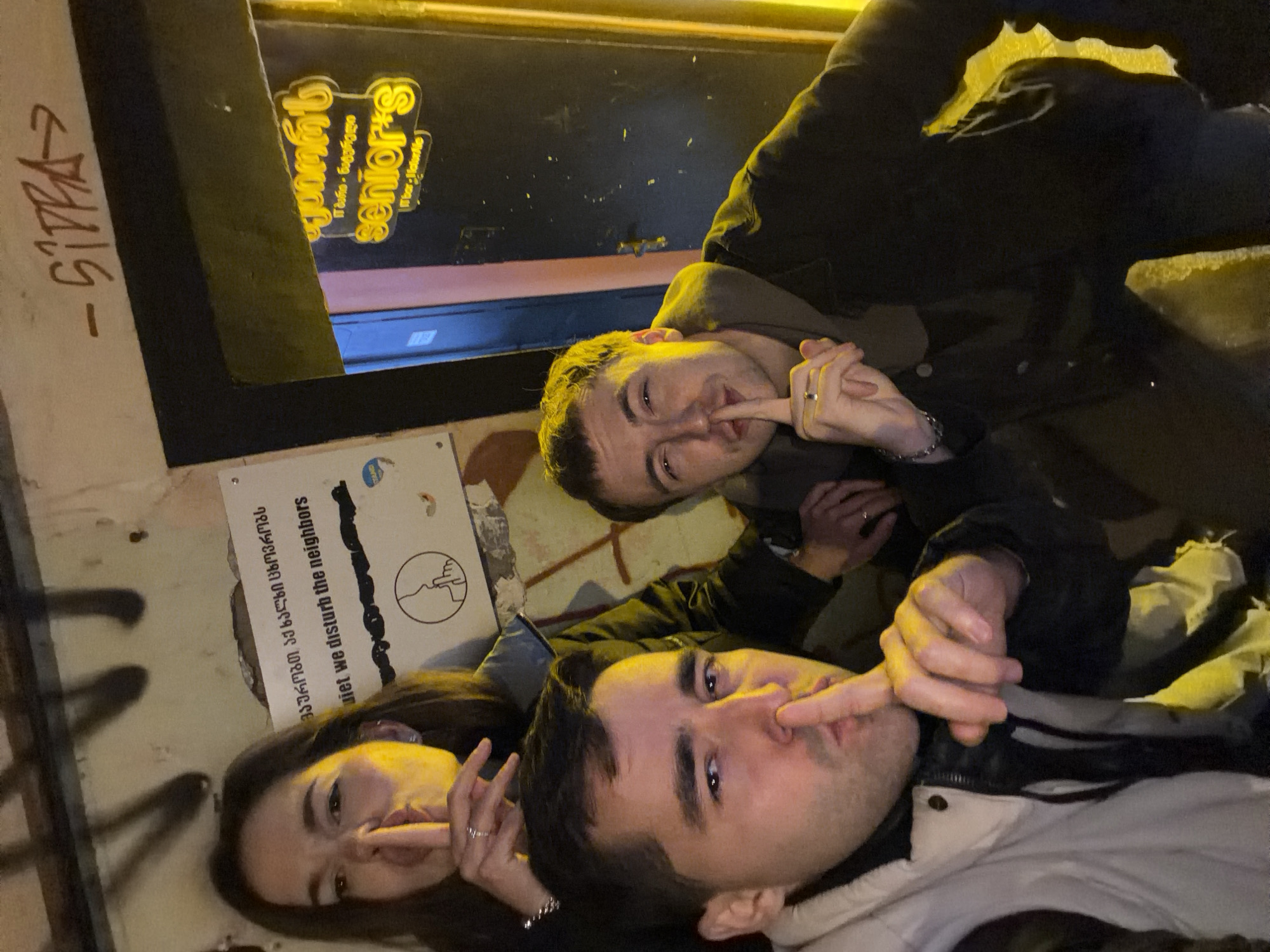 Three individuals are depicted in a low-light photograph taken at the entrance of "Senior's W. Bar - Wine / Cafe" in Tbilisi, Georgia. The establishment is identified by a yellow neon sign displaying "senior's" in English and Georgian script ("შენიორ'ს"), with "W. Bar - Wine / Cafe" and "Tbilisi, Georgia" also visible. Affixed to the textured wall to the left of the doorway is a white sign with text in Georgian ("გთხოვთ გაითვალისწინოთ") and English, reading "QUIET we disturb the neighbors," alongside a graphic depicting a face making a "shush" gesture.
All three individuals are actively performing the "shush" gesture by placing an index finger over their lips, an apparent acknowledgment of the sign. In the left foreground, a male with short dark hair, wearing a light-colored puffer vest over a dark top, looks directly at the camera with his right index finger on his lips. Behind him, a female with long dark hair, wearing a dark jacket, partially obscured, also looks towards the camera with her left index finger on her lips. A silver ring is visible on her left ring finger. To the right, a second male with short light brown hair, wearing a dark jacket over a hoodie, smiles faintly at the camera while making the "shush" gesture with his right index finger. He wears a silver ring on his right ring finger and a silver bracelet on his left wrist. The scene is illuminated by warm, yellow-orange artificial light, casting upward shadows. Graffiti, including black markings and a red inscription reading "-Sippa->", is visible on the wall above the sign and doorway.