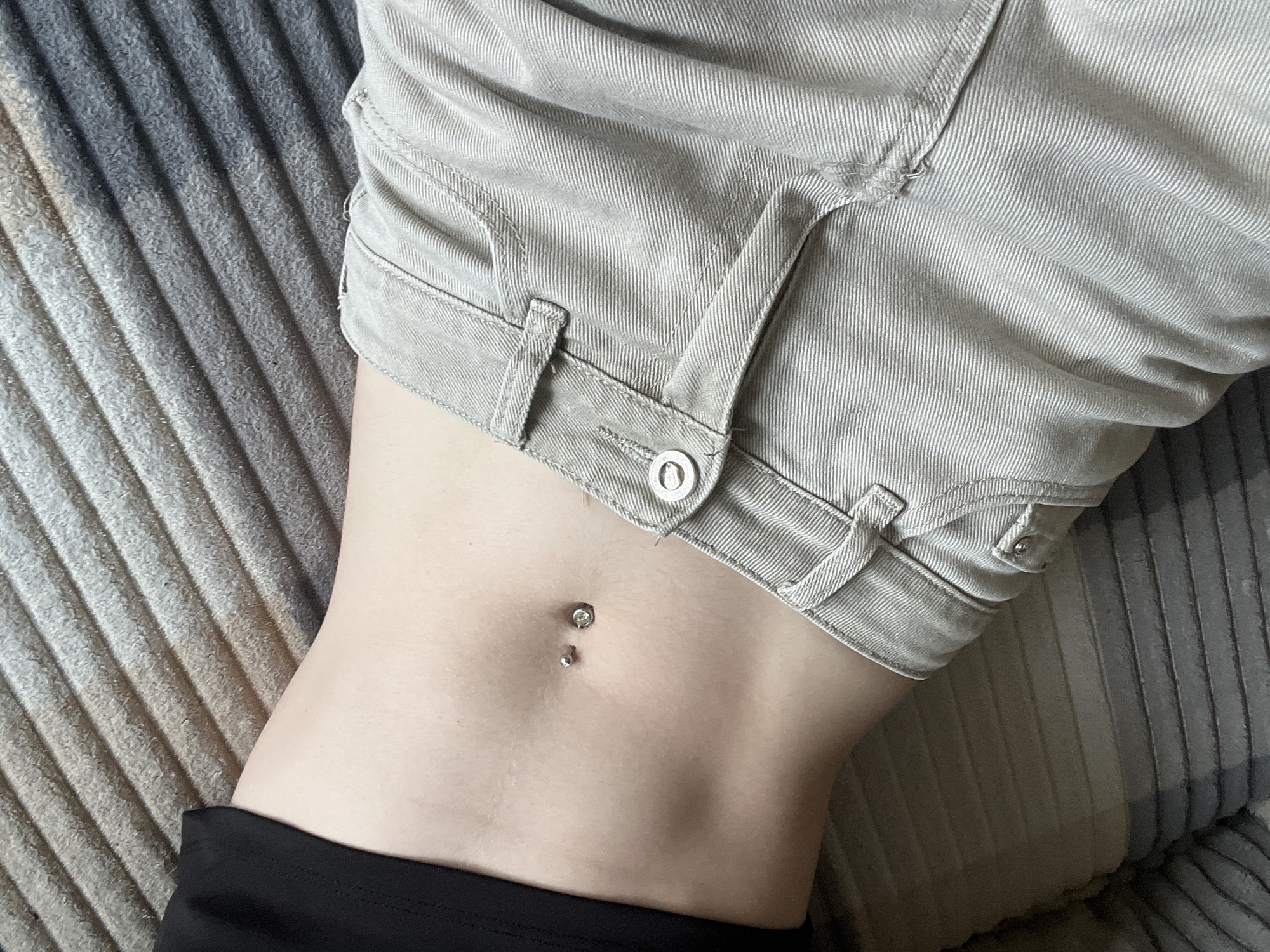 The image shows a close-up view of a person's midsection. The person is wearing light-colored denim shorts with a button closure. A navel piercing is visible. A black garment is visible below the waist. The background consists of a textured fabric. The location is unknown.