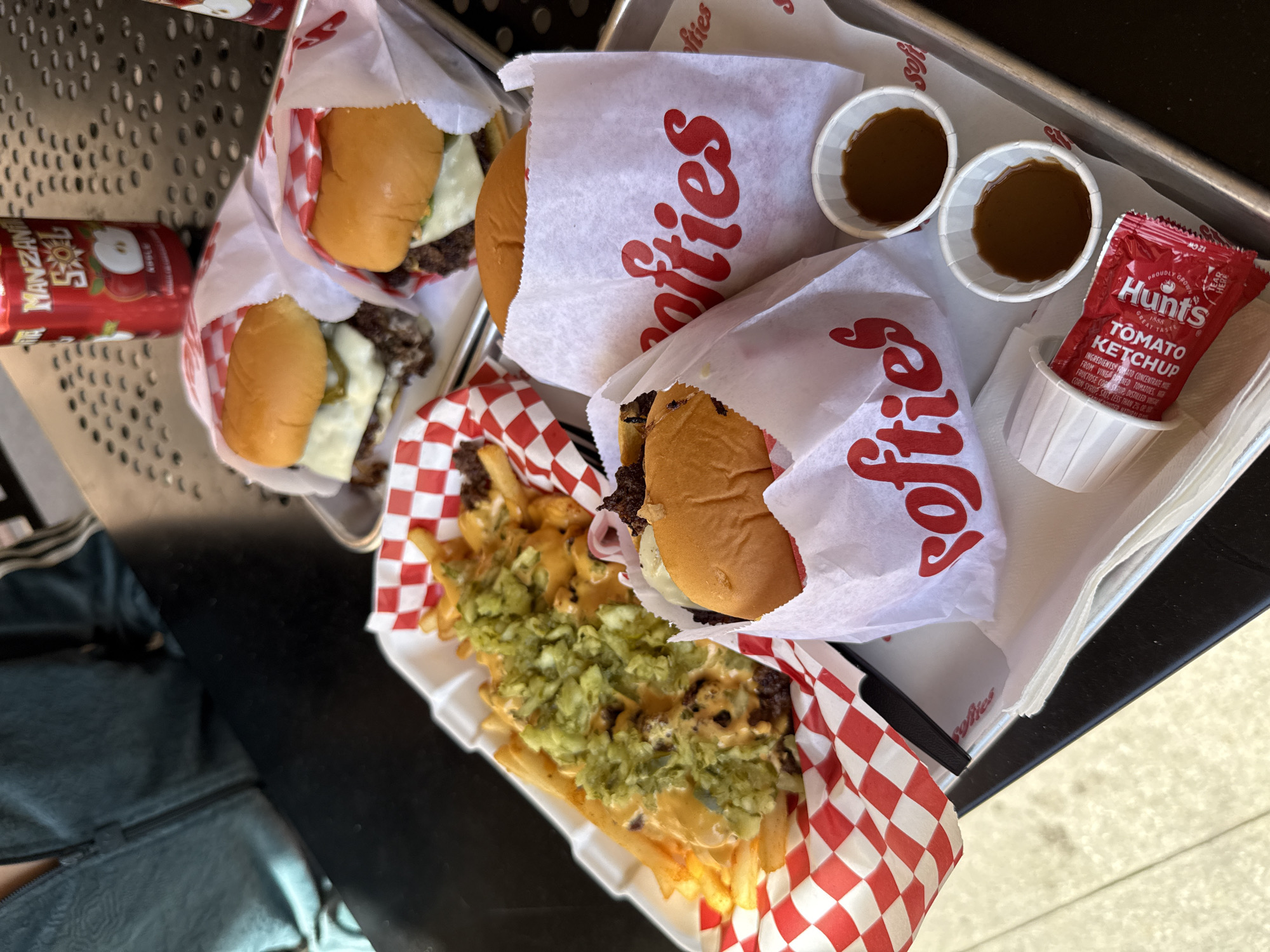 This media file captures a casual outdoor dining scene, likely in Los Angeles, United States of America, featuring an assortment of fast-food items. The main subjects are several burgers and a large serving of loaded fries, arranged on metal trays on a dark table.
At least five burgers, possibly sliders or smash burgers, are visible, wrapped in white paper featuring a prominent red "Softies" logo. Some burgers reveal melted cheese and meat patties. One substantial portion of golden fries is served in a white container lined with red and white checkered paper. These fries are generously topped with a creamy orange sauce and a vibrant green relish or chopped pickles.
Accompanying the meal are various condiments and drinks. Two small white cups are filled with a dark brown dipping sauce. A packet of Hunt's Tomato Ketchup is also present, with visible text including "Proudly Grown," "1888," "Great Taste," "Tomato Ketchup," and a partial ingredients list. A red aluminum can of "Manzana Sol" soda, featuring an apple logo, is partially visible.
The scene suggests a daytime setting due to the bright lighting and the light-colored ground visible in the background, hinting at an outdoor or semi-outdoor eating area. Part of a person's dark clothing with a zipper is seen on the lower left side, indicating someone is seated at the table, ready to enjoy the meal. The overall impression is one of a satisfying and hearty fast-food experience.