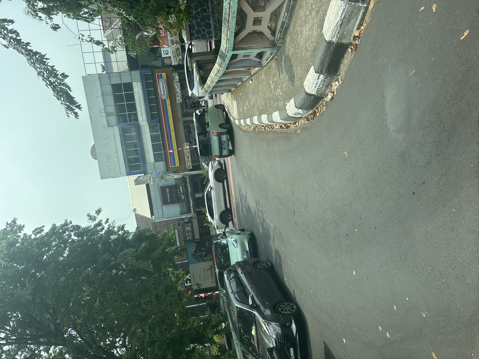 The image captures a street scene in North Jakarta, Indonesia, under a bright, overcast sky. The foreground shows a section of an asphalt road with a black and white striped curb and a concrete barrier. Several cars are present on the road, including a dark gray multi-purpose vehicle on the left, a light green compact car in the middle turning, and a white SUV further down the road. In the background, a row of buildings lines the street. One building features a prominent "Indomaret" convenience store sign with its signature red, yellow, and blue stripes. Other businesses with signage are also visible, suggesting a commercial area. Lush green trees with dense foliage overhang parts of the scene, particularly on the left side of the image. The overall impression is that of a typical urban environment with moderate traffic and commercial activity.