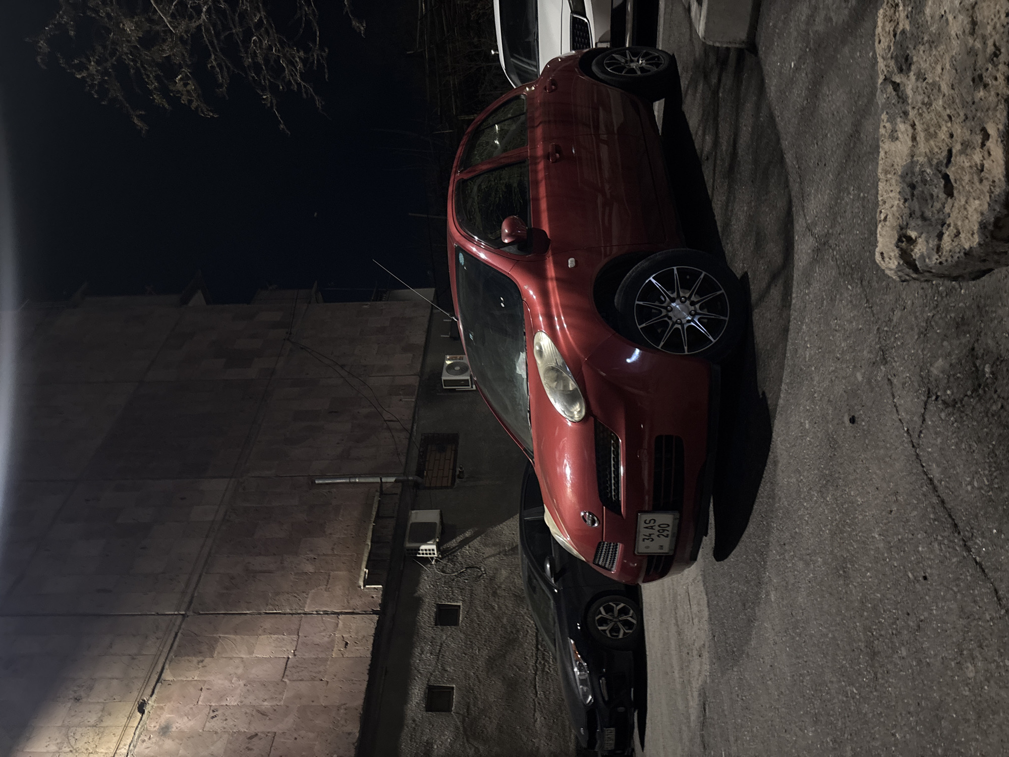 A red Nissan Micra is parked on a dark street in Yerevan, Armenia. The license plate reads "34 AS 290." The car is next to a stone wall on the right, and there is a tall building on the left. There is a dark-colored car partially visible below the red Micra. Air conditioning units are visible on the building to the left of the car.
