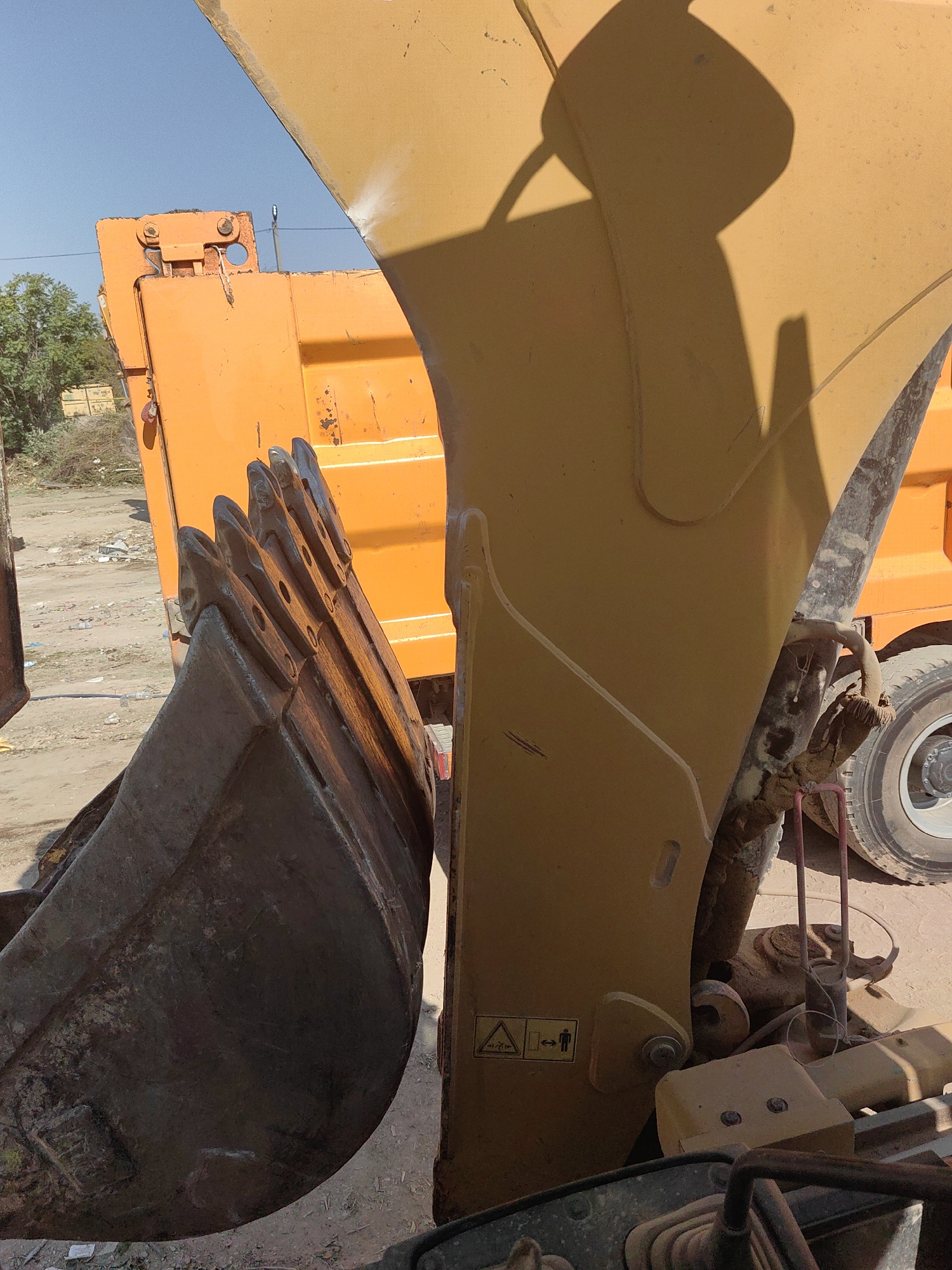 The image depicts a close-up view of heavy machinery, specifically the bucket of an excavator positioned next to the bed of an orange dump truck. The excavator bucket, made of weathered metal, is in the foreground, showing visible signs of wear and tear, including rust and dirt. It has a series of teeth along its leading edge. The dump truck's orange bed is visible behind the bucket, and a portion of its large tire and wheel assembly is also in view. The background shows a dusty, outdoor environment with scattered debris and vegetation. The sky is clear and blue. A shadow of the excavator's arm and bucket is cast across the yellow body of the excavator. There are no people visible in the image. The scene appears to be in an industrial or construction area, possibly a construction site or a yard where heavy equipment is stored or operated. The location is identified as Shymkent, Kazakhstan.