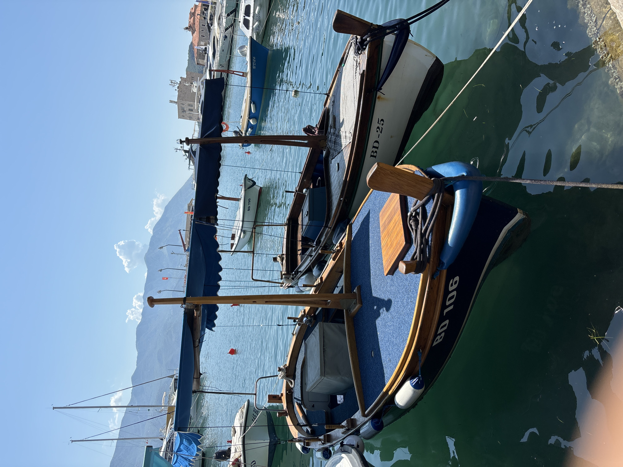 In Budva, Montenegro, two traditional wooden boats with blue awnings are moored in a clear blue harbor under a bright, sunny sky. The boat in the foreground, marked with "BD 106", has a blue hull and a wooden deck covered with blue carpet. A light-colored cooler is placed on the deck. The second boat, marked "BD-25", is positioned behind the first and appears to be white with a dark trim and a wooden interior. Several other boats, including yachts and smaller vessels, are visible in the background, along with buildings and a mountainous landscape. The water is calm, reflecting the blue sky and the surrounding structures.