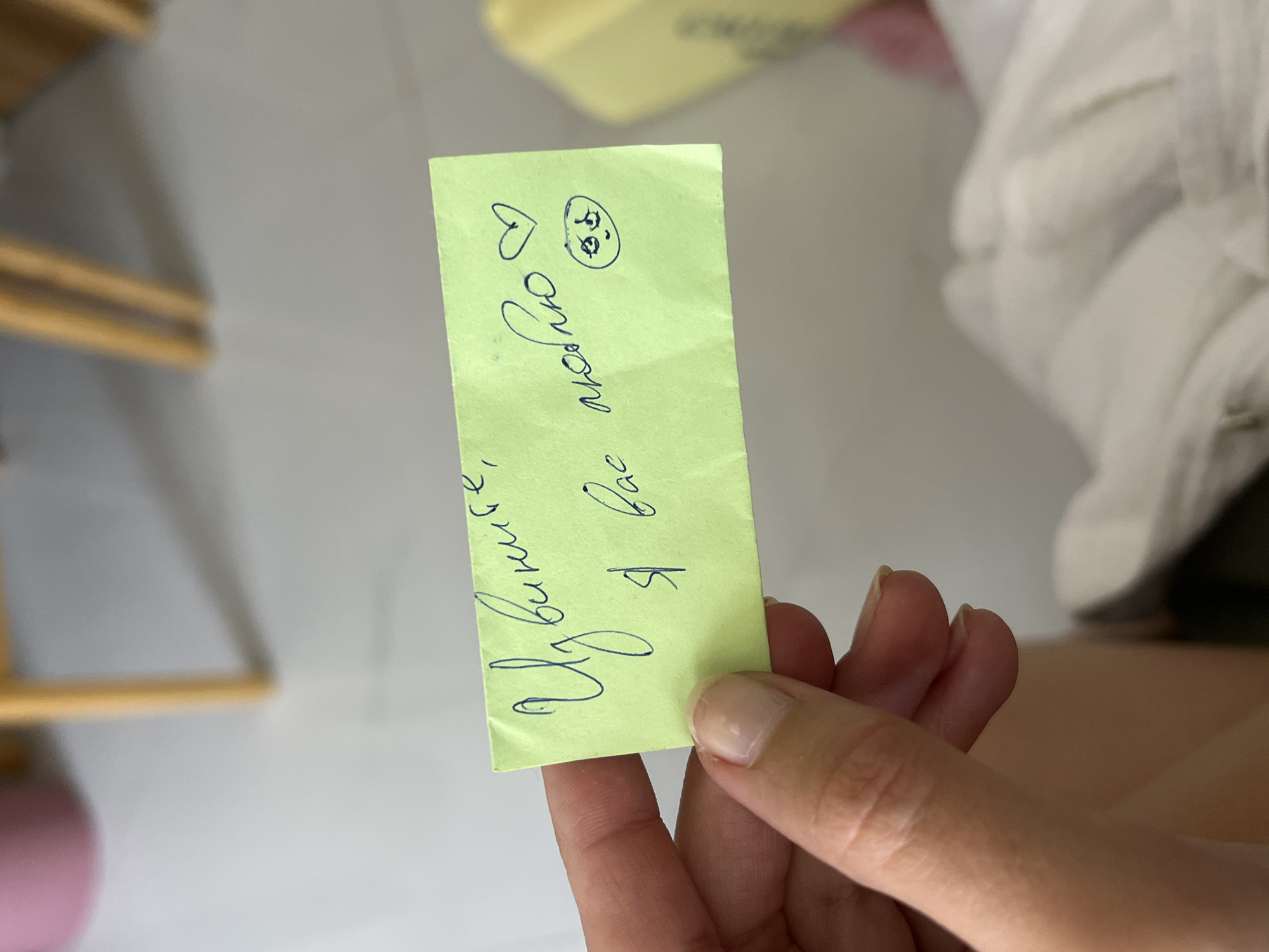A human hand with light skin tone and natural fingernails holds a rectangular, lime green note card, approximately 5x10 cm, in the foreground. The note displays handwritten text in dark blue ink, which translates from Russian as "Извините, я вас люблю" ("Sorry, I love you"). To the right of the text, a small heart symbol and a hand-drawn smiley face with wide eyes are present. The hand is positioned centrally, presenting the note towards the viewer. The background is blurred, showing an indoor environment with a light-colored surface, possibly a floor or table. Partially visible furniture includes blurred wooden legs, a pink object, and a yellow object. White, crumpled fabric is visible in the lower right. The scene is located in Nha Trang, Vietnam.