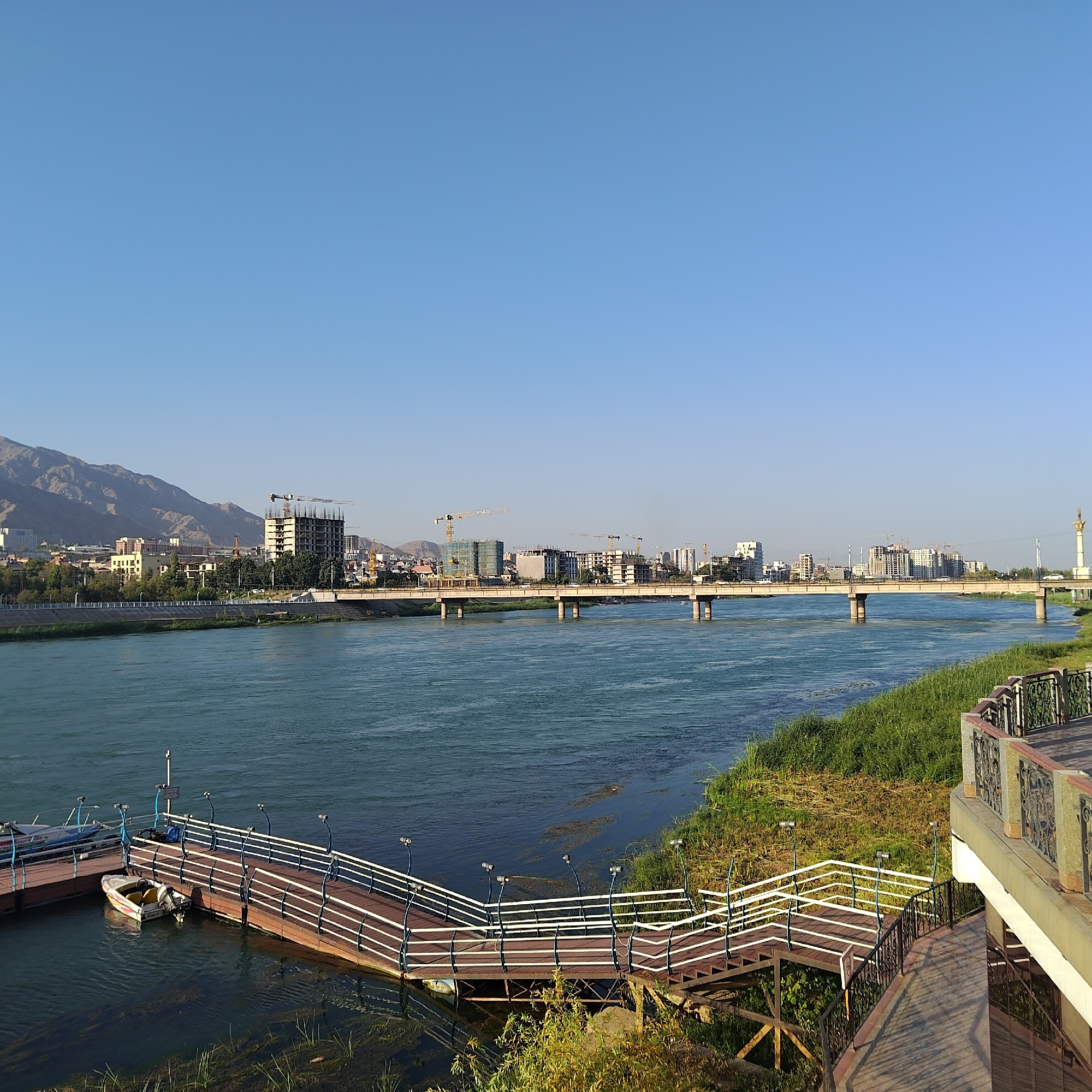 This is a wide-angle, eye-level outdoor shot capturing a serene river scene in Khujand, Tajikistan, likely on the Syr Darya river. The sky is a clear, cloudless blue, dominating the upper half of the frame. A wide river with turquoise-colored water flows horizontally across the middle ground. On the left bank of the river, a complex of buildings is visible, featuring a mix of modern structures, some under construction with visible cranes, and older, more traditional architecture. A mountain range forms the backdrop to the city. A concrete bridge with multiple spans crosses the river. The right bank of the river is characterized by lush green grass and vegetation. In the foreground, a wooden pier or walkway with white railings leads down to the water. A small, white motorboat is docked at the pier. Further to the right, a paved walkway with decorative railings and planters offers a vantage point overlooking the river and pier. The overall impression is one of a developed urban area adjacent to a natural river landscape, with evidence of ongoing construction.