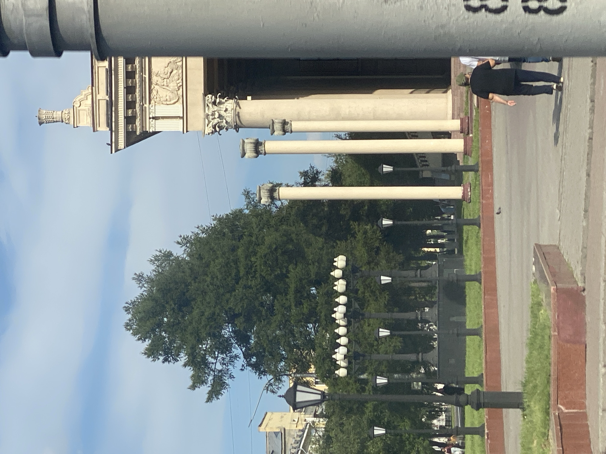 The image depicts an outdoor scene featuring a large building with classical architectural elements, including columns and decorative reliefs, on the right side. In the foreground, there are several lampposts and a paved walkway. A person wearing a dark shirt and pants is walking away from the camera. In the background, there are trees and a blue sky with some clouds. The location is identified as Novokuznetsk, Russia.