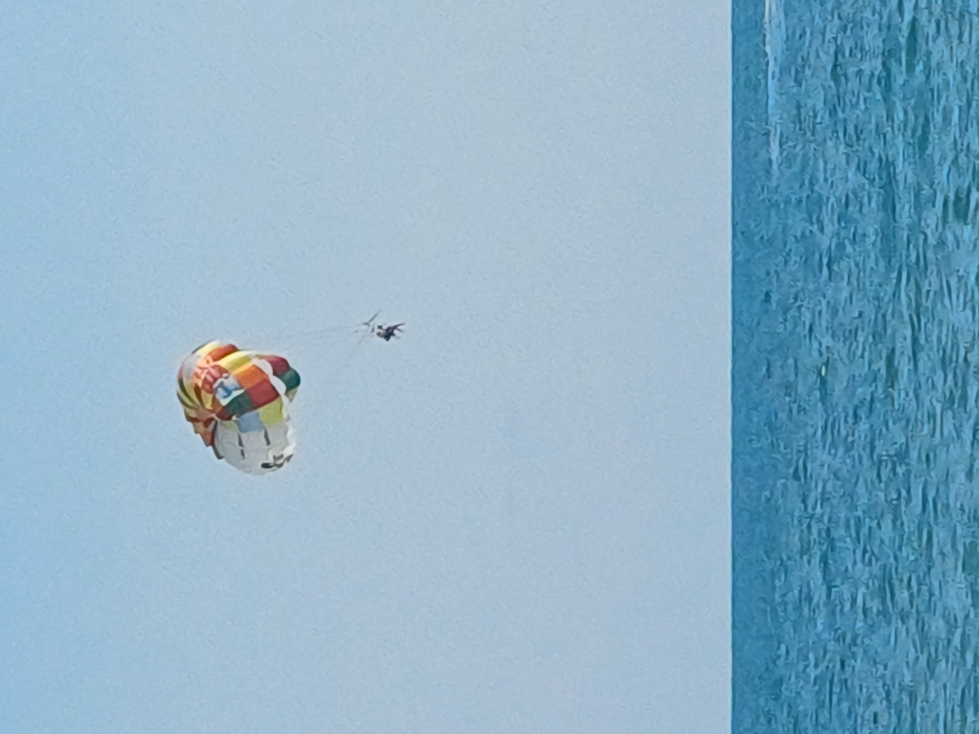 A parasail with a colorful canopy, featuring a patchwork of red, yellow, green, blue, and white segments, is visible against a pale blue sky. A single person is suspended beneath the canopy, appearing to be seated in a harness. The parasail is positioned above a body of water with gentle waves, likely the Black Sea, which occupies the lower portion of the image. The horizon line is clearly defined, separating the sky and the sea. The scene suggests an activity of parasailing taking place in a coastal area, possibly a resort destination like Sochi, Russia, though no specific landmarks or city details are discernible in the image itself.