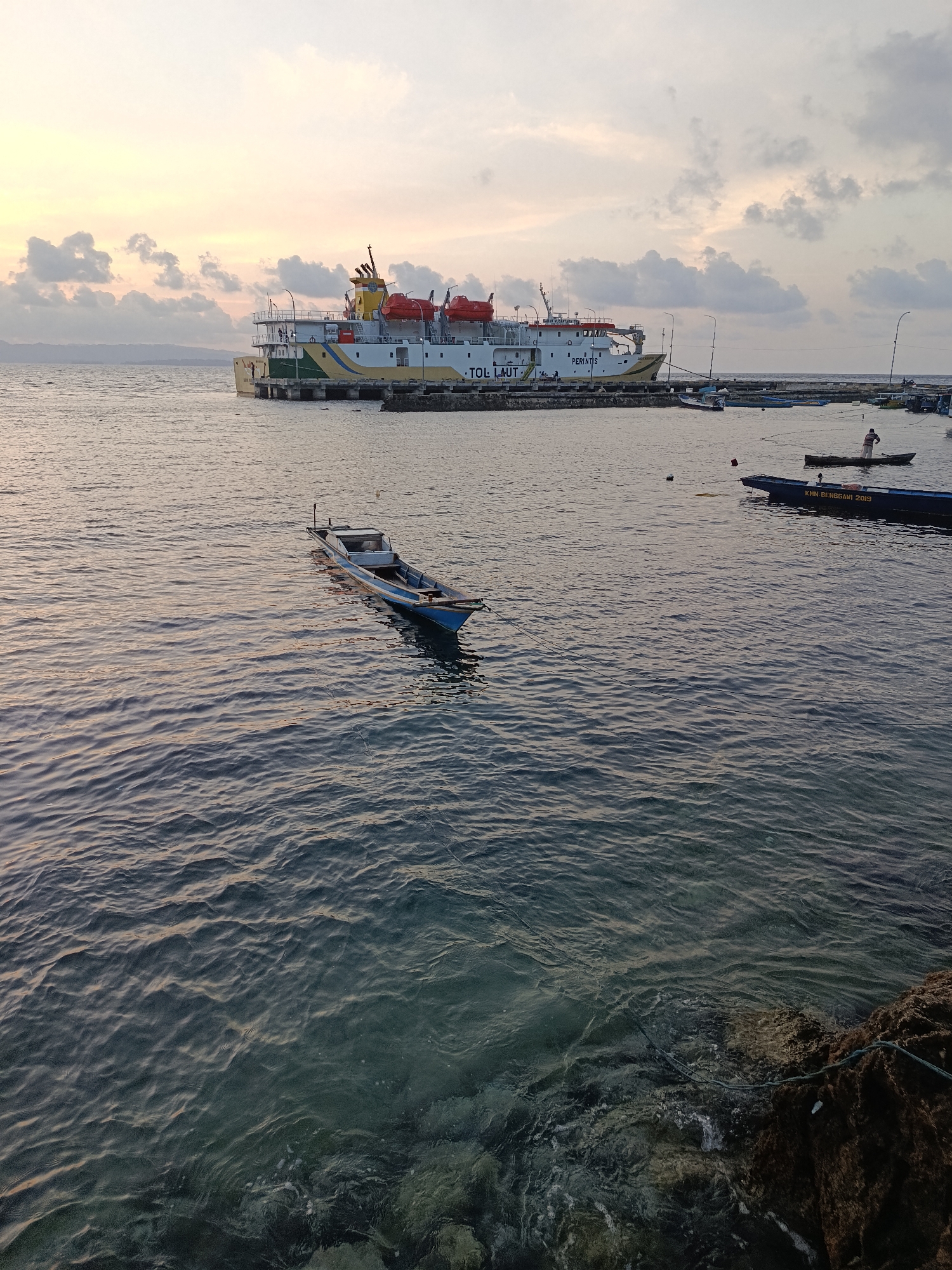 This image captures a tranquil scene at dusk in the waters off Mansalean, Indonesia. A large ferry, labeled "TOL LAUT," is docked at a pier in the background. Several smaller boats are scattered across the water. In the foreground, a blue wooden boat floats gently, its oars resting inside. The water is calm with gentle ripples, reflecting the soft hues of the sunset sky, which is adorned with scattered clouds. The shoreline is visible with some rocks and vegetation. One person is visible in a small boat in the distance, seemingly engaged in fishing.