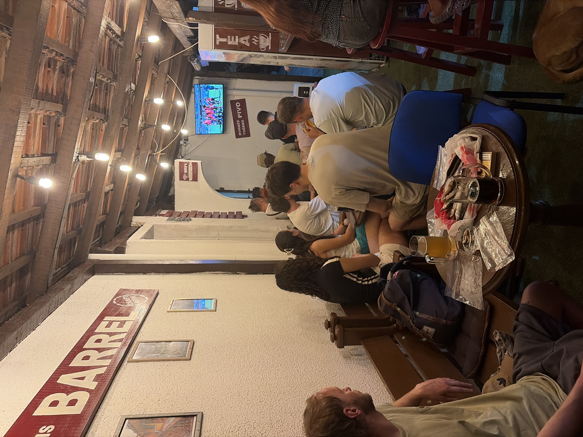 The image depicts an indoor scene at a bar in Bar, Montenegro. The bar has a rustic ambiance with a wooden beamed ceiling and exposed brick accents. A large sign above the seating area reads "BARREL". Several people are seated at tables and on benches, engaged in conversation and watching a television screen that is displaying a sports match. The tables are laden with drinks, including glasses of beer, and there are also ashtrays and cigarette packs visible. The lighting is dim, with string lights hanging from the ceiling, contributing to the relaxed and casual atmosphere of the establishment.