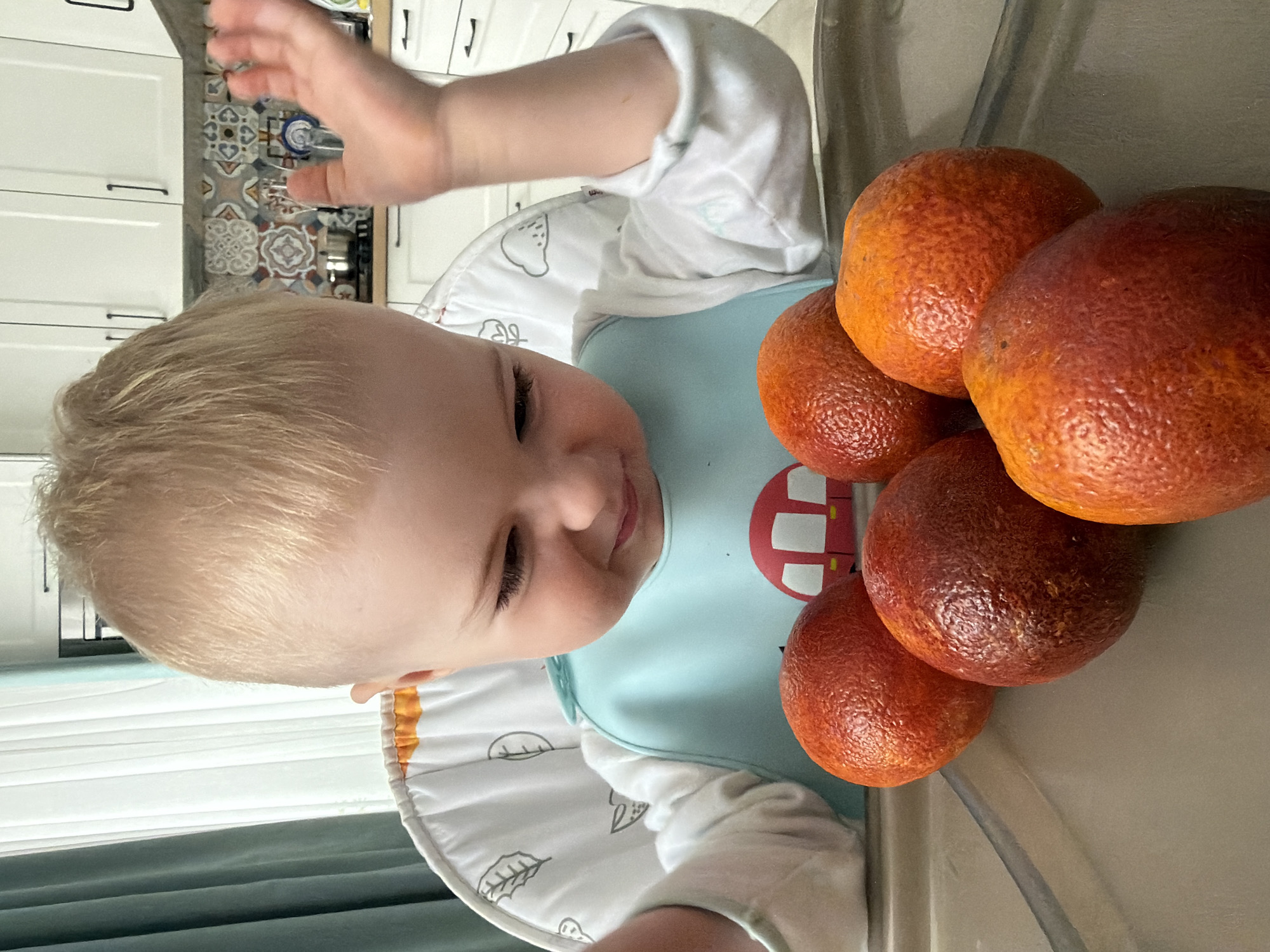 A baby sits in a high chair with five blood oranges on the tray. The baby has blonde hair, fair skin, and is wearing a bib and a white long-sleeved shirt. The kitchen background is visible with white cabinets and a patterned backsplash. The scene is indoors, likely in a home. There is no indication of the location being Votkinsk, Russia.