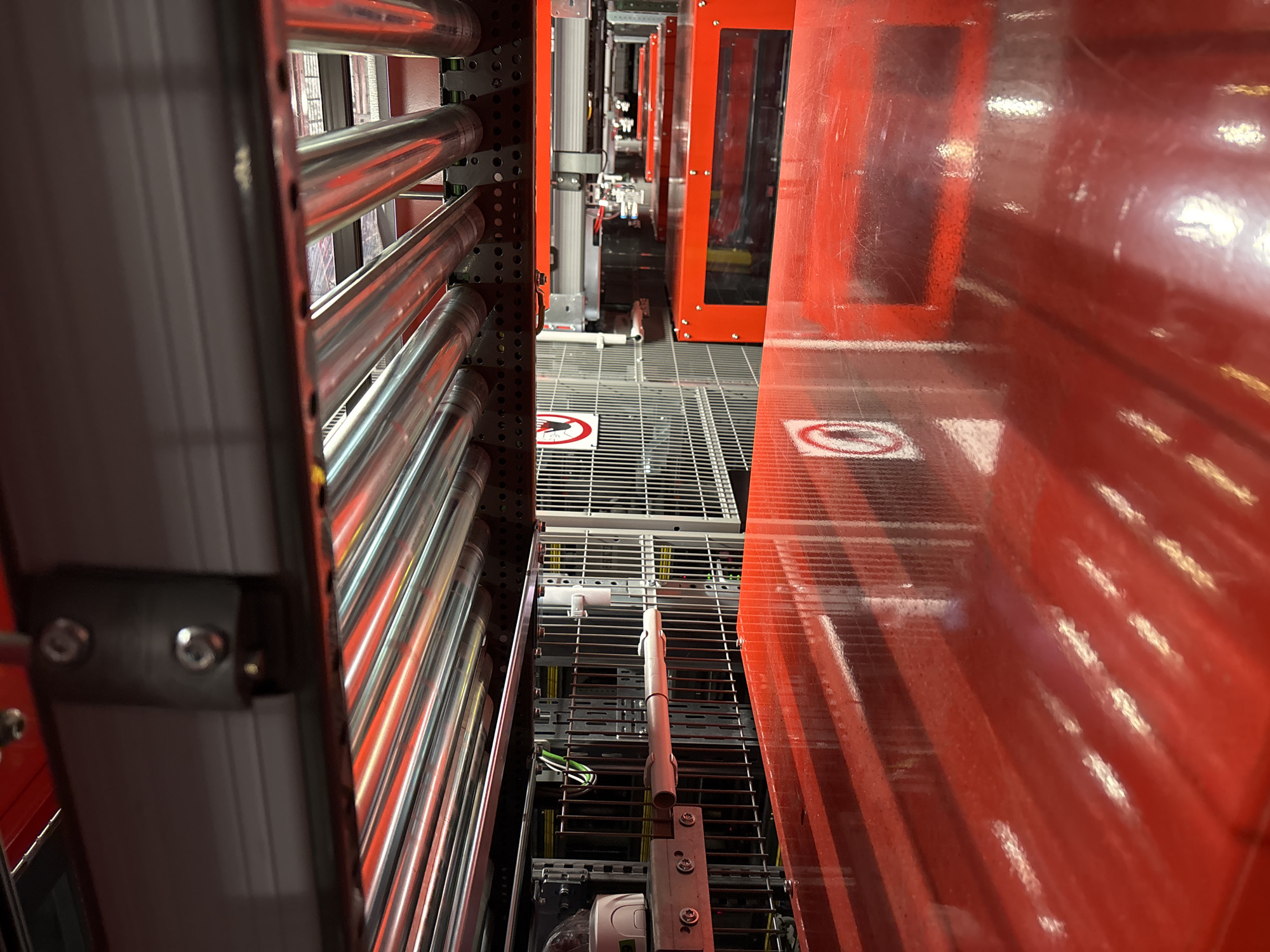 The image captures a low-angle perspective of a conveyor system, likely within an industrial or logistics facility. The foreground is dominated by a glossy, bright red surface, possibly a conveyor belt or a transfer table, with reflections of the overhead structures. Above this red surface, a series of parallel, metallic roller conveyor sections are visible, running horizontally. These rollers are illuminated by red lights, creating a warm, almost fiery glow.
In the mid-ground, behind the roller conveyor, a silver-colored metal grid structure is apparent, suggesting partitions or safety fencing. Attached to this grid are various components, including what appear to be sensors or cameras, along with some electrical wiring. A white cylindrical object is also mounted on the grid. Further in the background, more of the red conveyor system can be seen, with a red frame enclosing a transparent panel, hinting at automated machinery or processing equipment. Two circular warning signs with a red border and a diagonal line through them are visible on the metal grid, one in the left mid-ground and another on the red structure in the right mid-ground, suggesting a safety or operational instruction.
The environment appears to be a clean, well-lit industrial setting. The overall impression is one of advanced automation and efficient material handling. There are no people visible in the image.
This scene is located within the city of Mannheim, Germany. Specifically, it is inside a facility operated by DHL, a prominent logistics company, likely at one of their parcel sorting centers or distribution hubs. The specific location within Mannheim could be one of their operational sites, contributing to the flow of goods within the city and beyond.