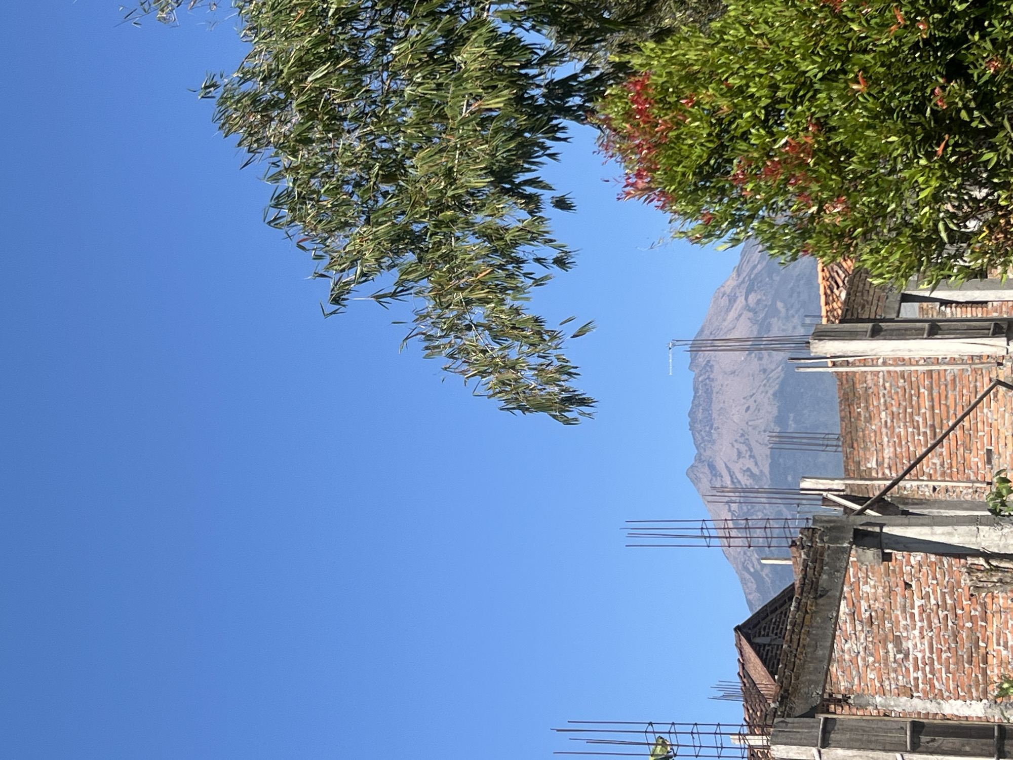 In Danupayan, Indonesia, a landscape photograph captures a clear blue sky above an unfinished brick structure with exposed rebar. A mountain, likely a volcano, dominates the background, its slopes appearing rugged and possibly volcanic ash-covered. In the foreground, lush green trees with some red foliage partially obscure the scene, with one tree's branches hanging down from the upper right corner. The construction site suggests ongoing development, contrasting with the natural grandeur of the distant mountain.