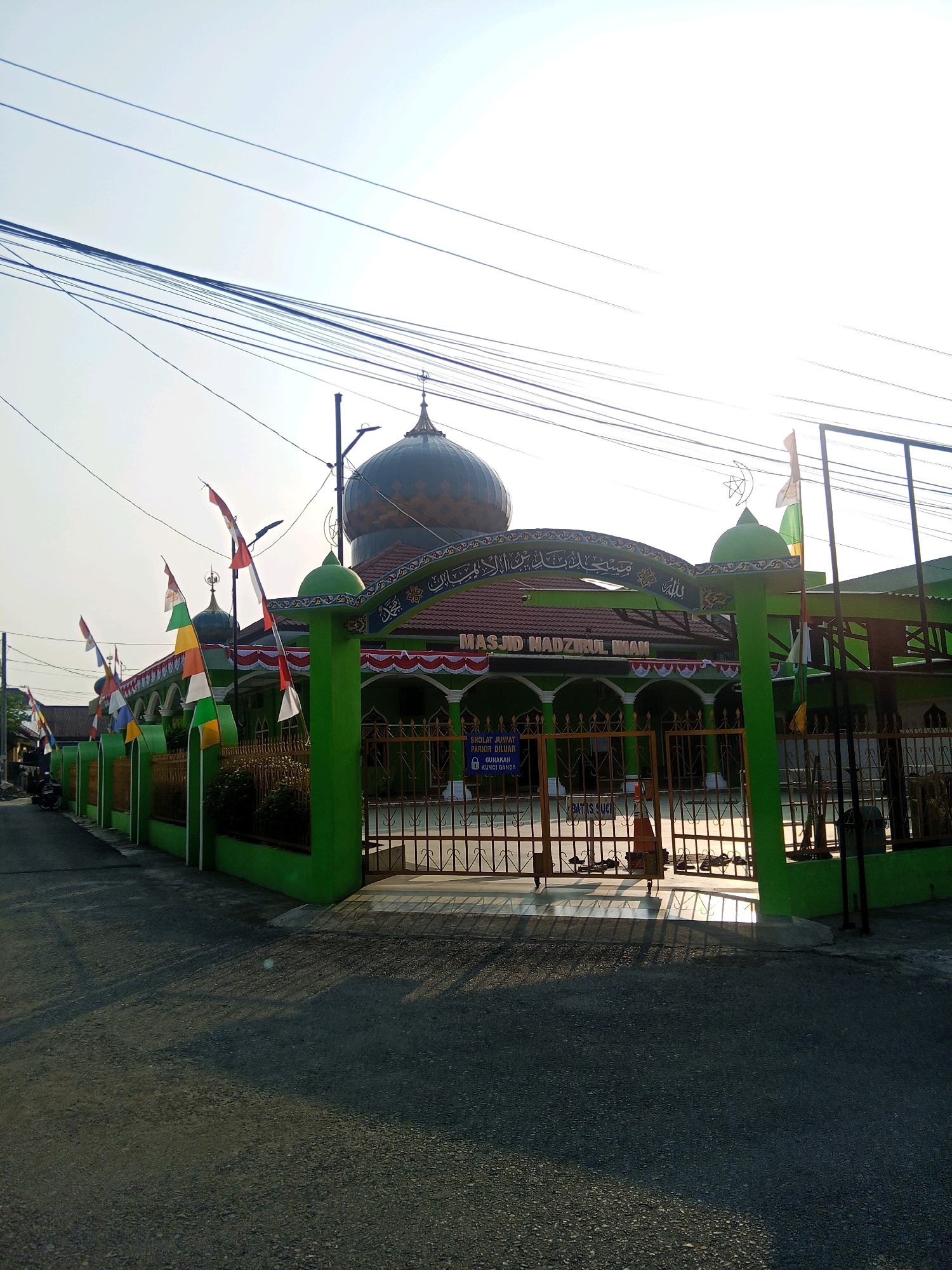 The image shows the front of the Nadzirul Iman Mosque in Lubuklinggau, Indonesia, under a bright, hazy sky. The mosque is painted green and features a large dome. Colorful Indonesian flags are displayed along the fence. The mosque entrance is a metal gate, and a sign reads "SHOLAT JUMAT PARKIR DILUAR GUNAKAN KUNCI GANDA." The ground in front of the mosque is paved, and shadows from the fence and flags are cast across it. A few motorcycles are parked to the left of the mosque. The overall scene suggests a religious site in a residential or semi-urban area of the city.