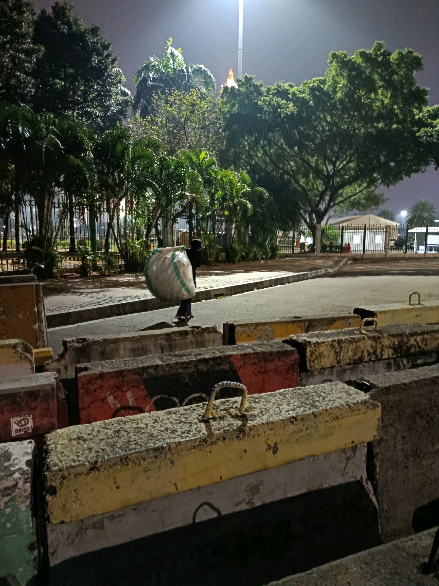 A person walks across a paved area at night, carrying a large, white sack over their shoulder. The sack has green stripes. The person is wearing a dark hooded garment and dark pants. In the foreground, there are several large concrete barriers, some painted with red, yellow, and black markings. Behind the person, there is a row of palm trees and other foliage, with a fence and a building visible in the background. A bright light illuminates the scene from above, likely a streetlamp or floodlight. The overall environment appears to be an outdoor public space, possibly a park or a street adjacent to a park, located in Central Jakarta, Indonesia.