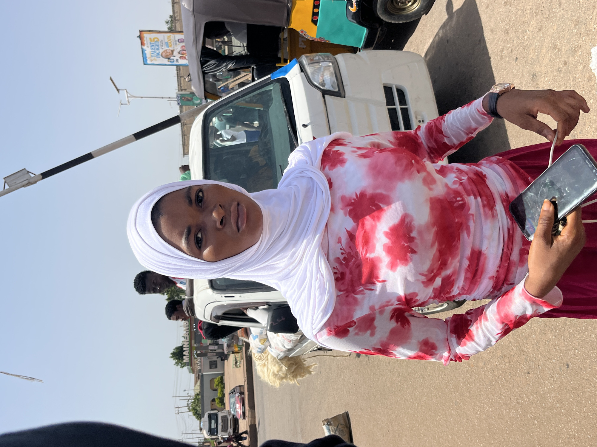 In an outdoor, urban setting, a young woman wearing a white hijab and a white and red tie-dye patterned long-sleeved shirt stands in the foreground, holding a smartphone. Her left arm is slightly bent, and her fingers are interacting with a charging cable connected to the phone. She is also wearing a dark pink skirt. Behind her, a white flatbed truck is visible, loaded with what appears to be textiles or some form of cargo, with several men standing in the truck bed. To the right of the truck, a yellow and green auto-rickshaw is parked, with a driver and passenger inside. In the background, there are buildings, including one with a "MAINLAND" sign, and other vehicles, suggesting a busy street or market area. The sky is clear and blue. The overall scene depicts a moment of everyday life on a street.