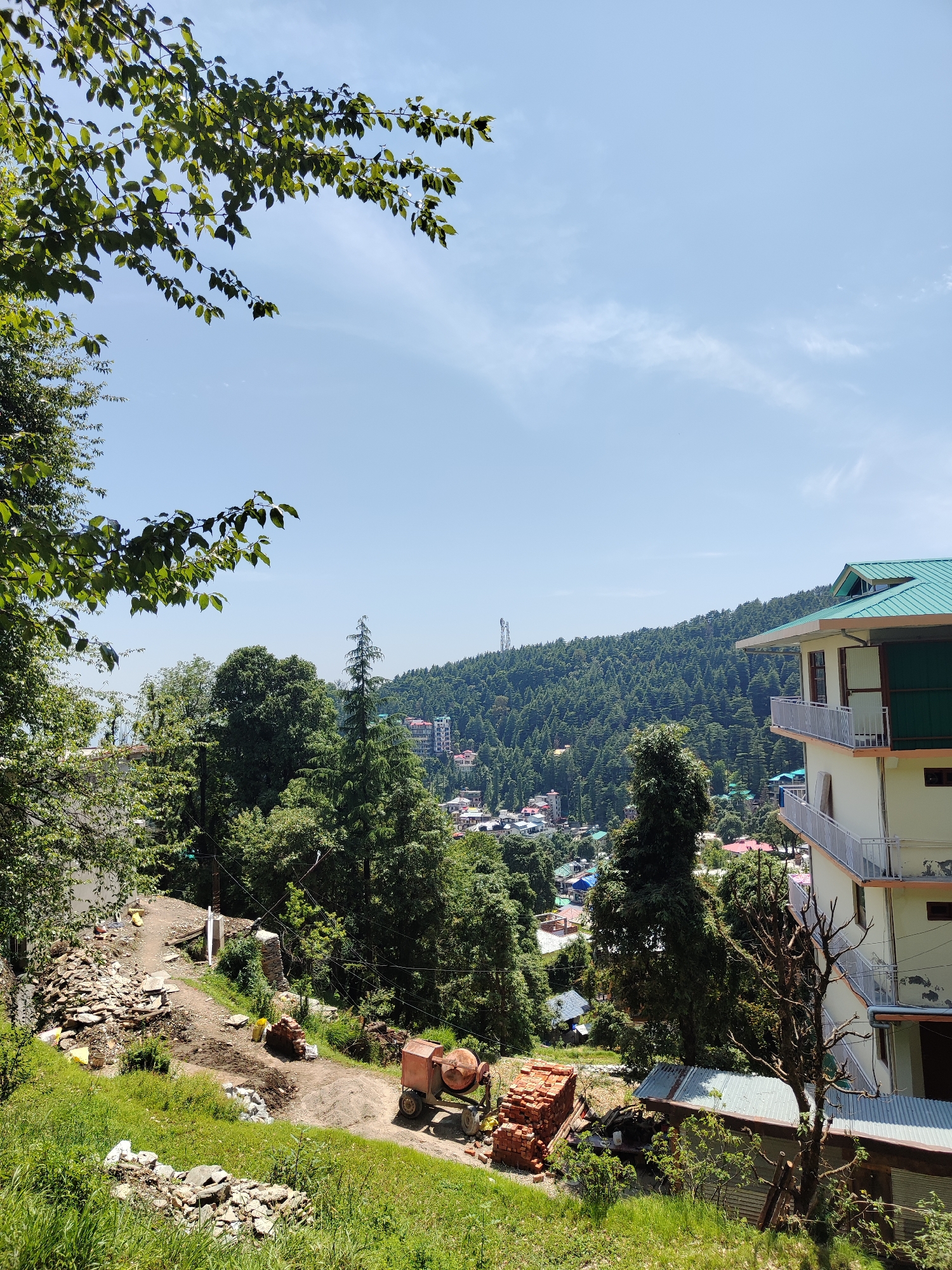 The image depicts a view in Haini, India, showcasing a landscape with a blend of natural and built environments. In the foreground, there is a partially constructed area with building materials such as bricks, sand, and a cement mixer. Vegetation includes green grass, trees, and shrubs. A narrow dirt path leads towards a distant view of the town. In the midground, there is dense foliage and buildings scattered amongst the trees. The background reveals a hillside covered with trees, with residential buildings visible. In the distance, a tower-like structure can be seen on the crest of a hill. The sky is blue with light cloud cover. A multi-story building with balconies is to the right of the image.