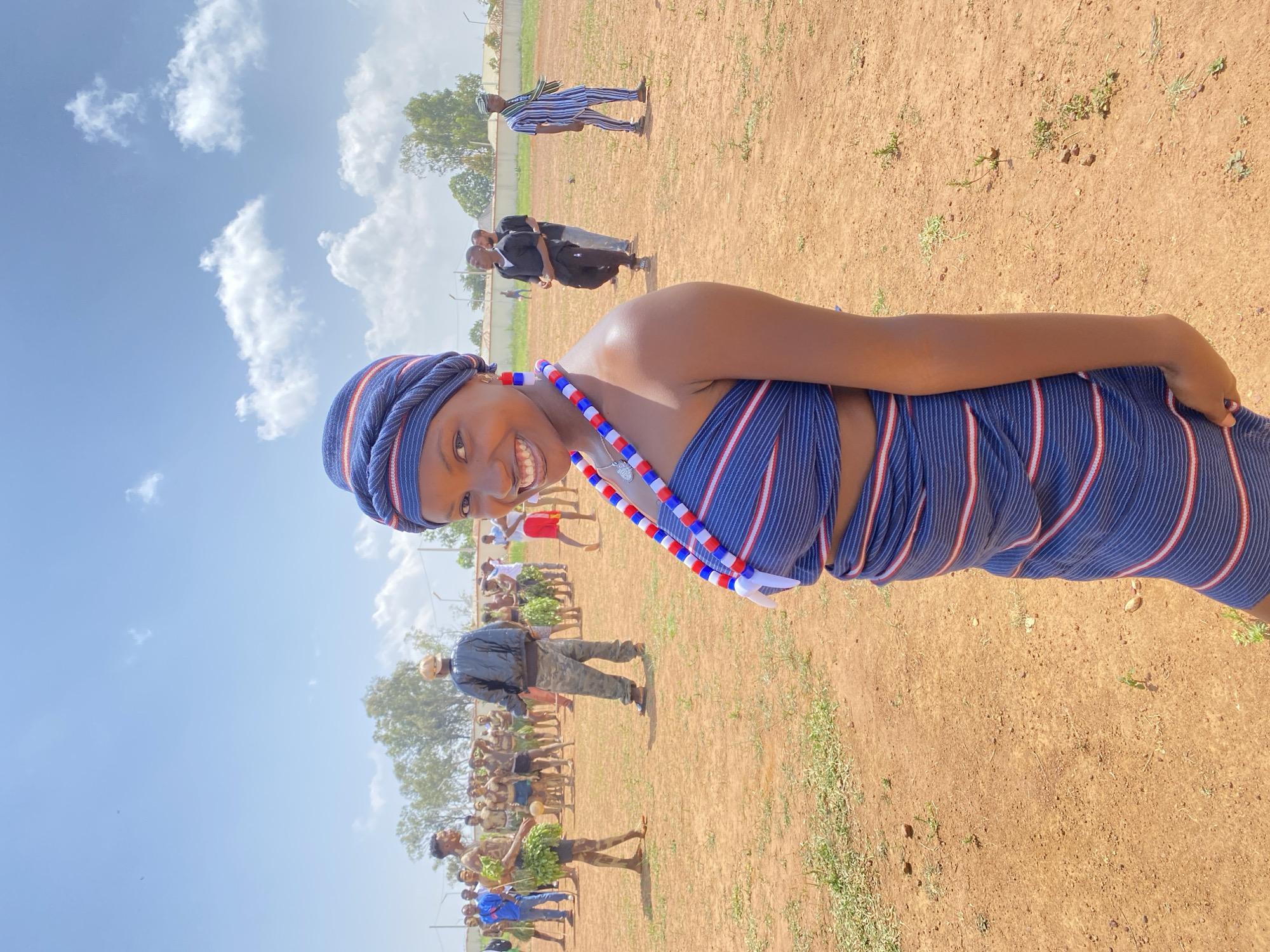 A smiling dark-skinned woman stands in a dirt field in Durimi, Nigeria, wearing a blue and white striped traditional garment and a beaded necklace. Behind her are several groups of people, one group dressed in suits, and another wearing what appears to be traditional garb. Trees and buildings are visible in the background under a sky with scattered clouds.