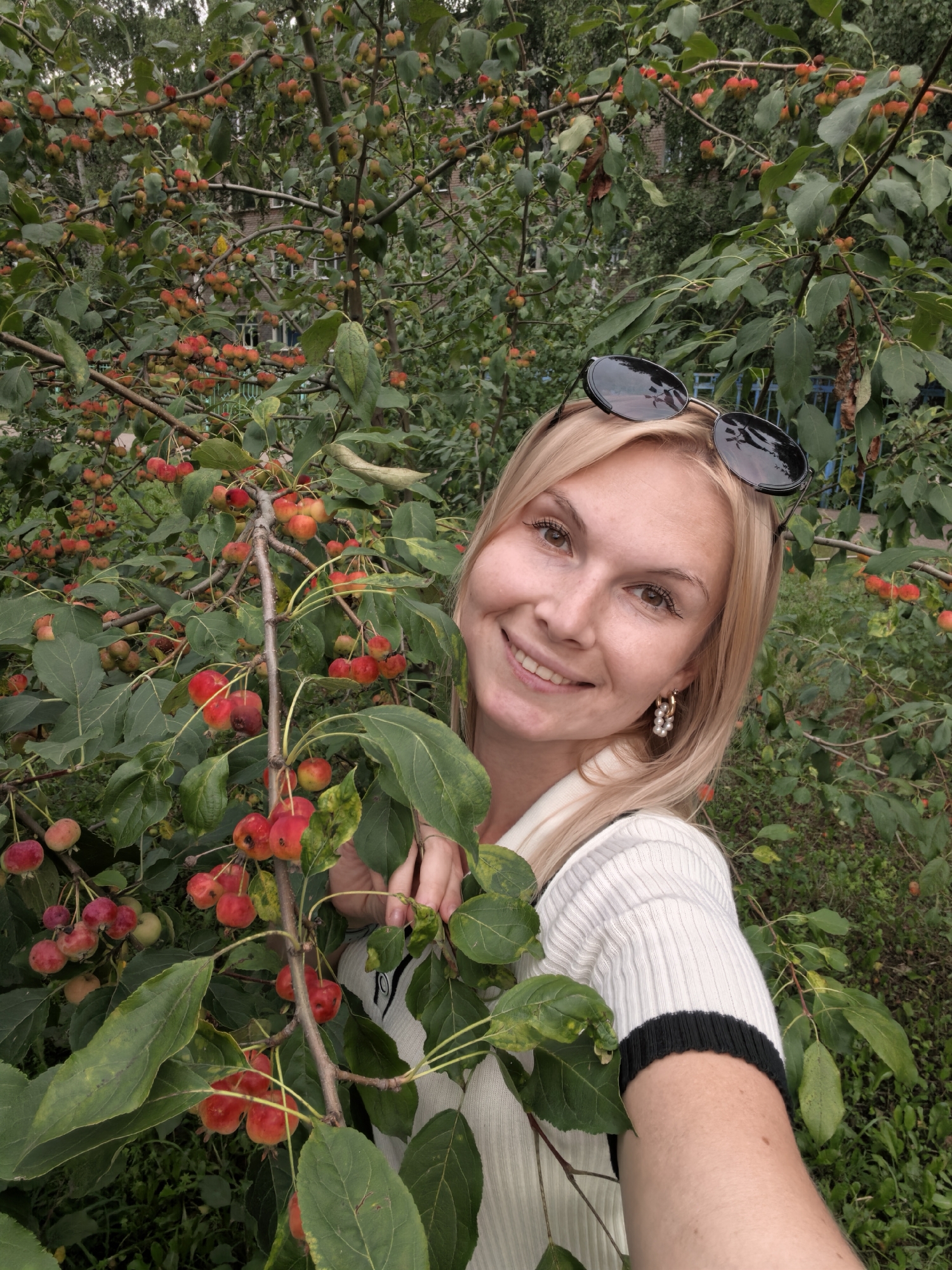 The image shows a blonde woman with sunglasses on her head, wearing a white top with black trim. She is smiling and looking at the camera, holding a branch with small red fruits in her right hand. She is standing outdoors in front of a tree laden with similar fruits. The background shows more trees and some buildings, suggesting an urban or suburban environment. The lighting indicates it is daytime.