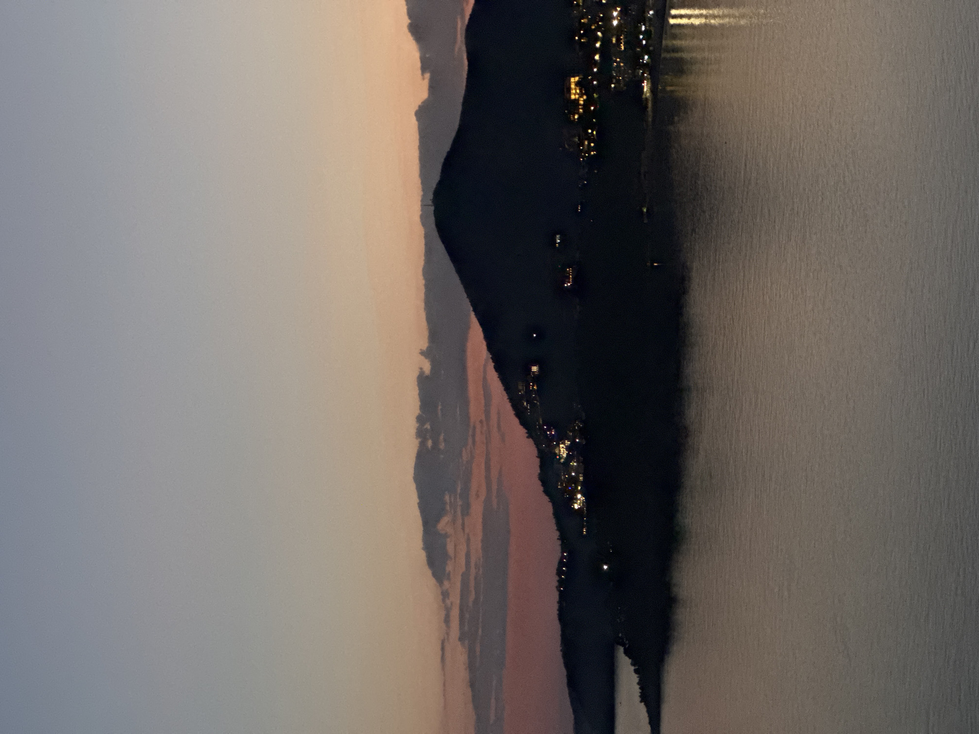 The image shows a coastal landscape at dusk or dawn. The sky transitions from a light gray on the left to a peachy orange towards the center. A dark, silhouetted mountain range or land mass divides the sky from a body of water on the right. Lights from buildings are visible along the coastline, reflecting slightly in the water. The scene is likely Rafailovići, Montenegro, based on the coastal setting, terrain, and architectural features. The image appears to be rotated 90 degrees clockwise.