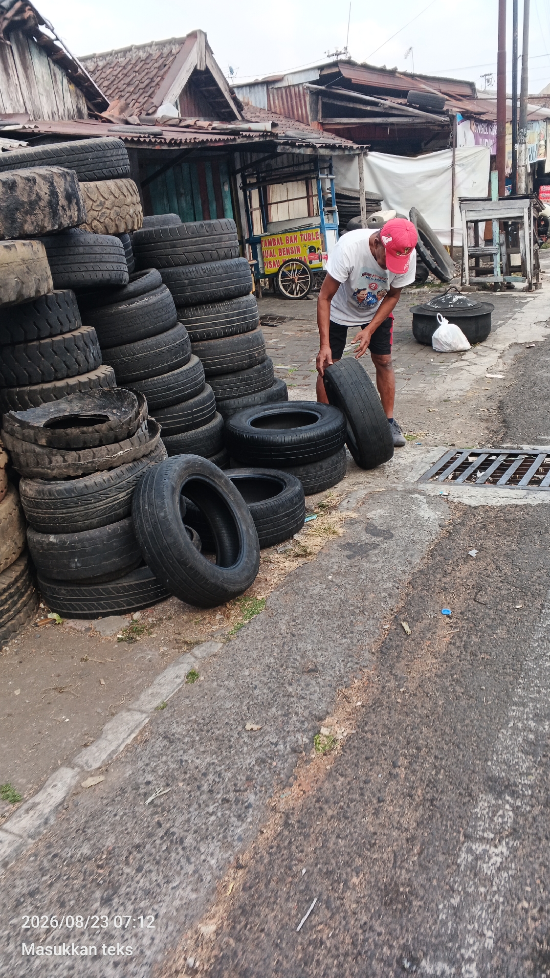The image depicts an outdoor scene in the city of Malang, Indonesia. In the foreground, a man wearing a white t-shirt with a graphic print and a red baseball cap is interacting with a stack of used tires. He is holding one tire, appearing to be either placing it or retrieving it from the pile. The tires are stacked haphazardly on a paved surface, with some tires also lying on the ground.
Behind the man and the tires, there is a roadside stall or shop with a weathered wooden structure and a corrugated metal roof. A sign on the stall, partially visible, appears to advertise "TAMBAL BAN TUBL" and "JUAL BENSIN" (tire repair and gasoline sales). A bicycle with spoked wheels is leaning against the stall. Further in the background, there are more structures, possibly residential or commercial buildings, with zinc roofs. The environment suggests a typical street scene in a less developed urban area.
To the right of the man, there is a large black cauldron-like object and a white plastic bag on the ground. A metal grate is visible in the paved area near the man's feet. The ground is a mix of asphalt and dirt. The overall lighting suggests it might be late afternoon or early morning. The timestamp at the bottom left of the image reads "2026/08/23 07:12" followed by Indonesian text "Masukkan teks," which translates to "Enter text." This indicates the image was captured on August 23, 2026, at 7:12 AM.