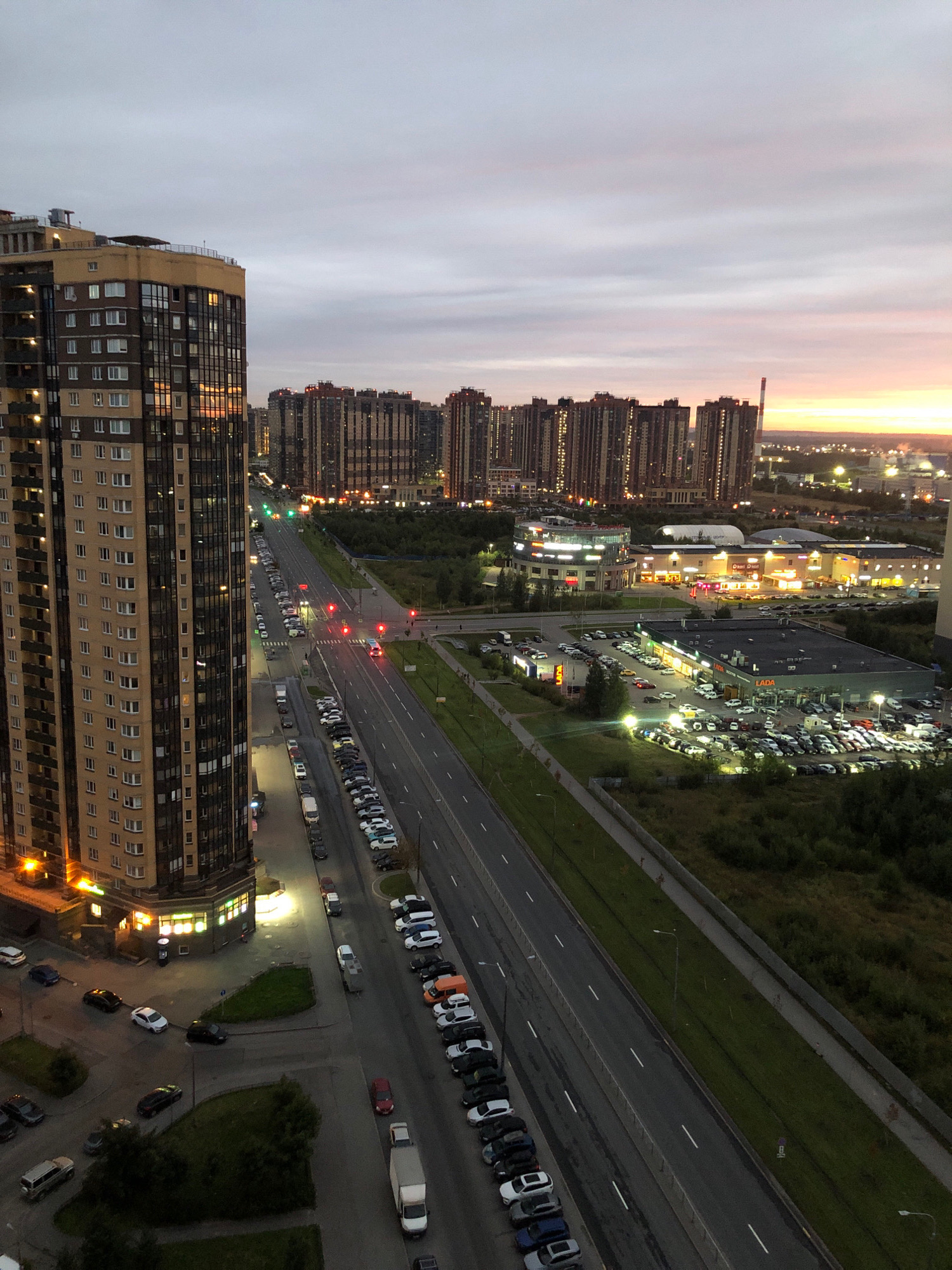 The image is a high-angle, vertical shot of a city street in Babino, Russia at dusk. A large, modern, multi-story apartment building with beige and dark paneling dominates the left side of the frame. The building has numerous windows and illuminated sections on its lower floors. Across the street, a long, multi-lane road stretches into the distance, lined with parked cars on both sides. Traffic lights are visible, some showing red and green. In the background, a series of identical, tall, dark residential buildings with illuminated windows form a dense cityscape. Further in the distance, a wider panorama of city lights and buildings is visible against a twilight sky with streaks of orange and pink from the setting sun. The overall scene depicts a suburban or outskirt area of a Russian city.
