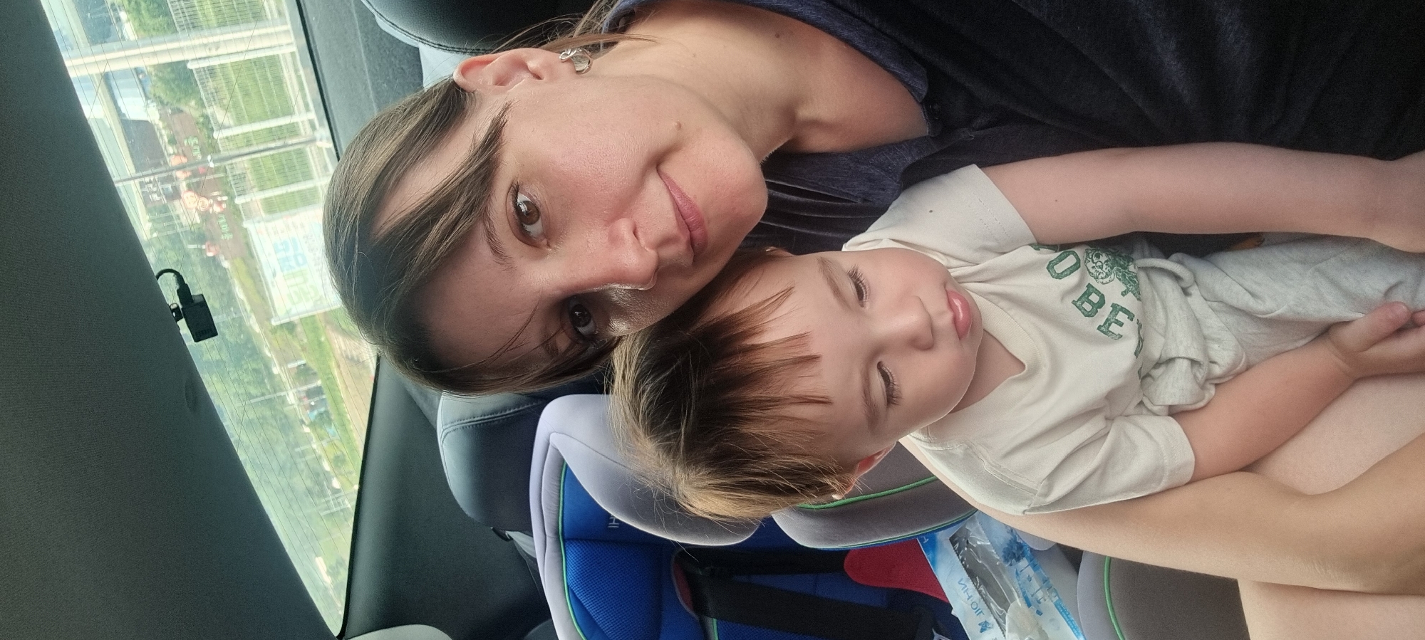 This image captures a tender moment between a woman and a young child inside a vehicle. The woman, positioned in the foreground, has brown hair and brown eyes and looks directly at the camera with a gentle smile. She is wearing a dark-colored top. Resting against her, a young child, likely a toddler, appears to be sleeping or very peaceful with eyes closed. The child has light brown hair and wears a light-colored t-shirt with green text, which clearly shows "OBE" (possibly part of a word like "GLOBE" or "ROBE").
The setting is the back seat of a car, with a blue and grey child safety seat partially visible. Through the car's rear window, a bright daytime outdoor scene is visible. The landscape outside appears to be rural or semi-urban, featuring green fields, some distant buildings, and what looks like infrastructure or roads. A small, black electronic device, possibly a dashcam or air freshener, hangs from the top edge of the rear window. The overall mood of the scene is calm and affectionate, suggesting a moment of rest or comfort during a car journey in Dunnae-myeon, South Korea.