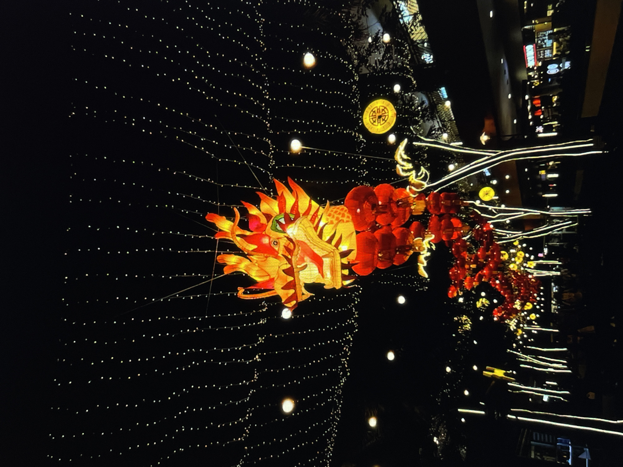 The image depicts a nighttime scene with a large, illuminated dragon lantern as the focal point. The dragon is predominantly red and yellow, with details of its head and body clearly visible. The lantern is suspended amidst a backdrop of strings of small, white lights, creating a shimmering effect. In the background, there are glimpses of urban architecture, including the edges of buildings and signs that suggest a city environment. The overall lighting is low, but the lanterns and city lights provide illumination, enhancing the festive atmosphere.