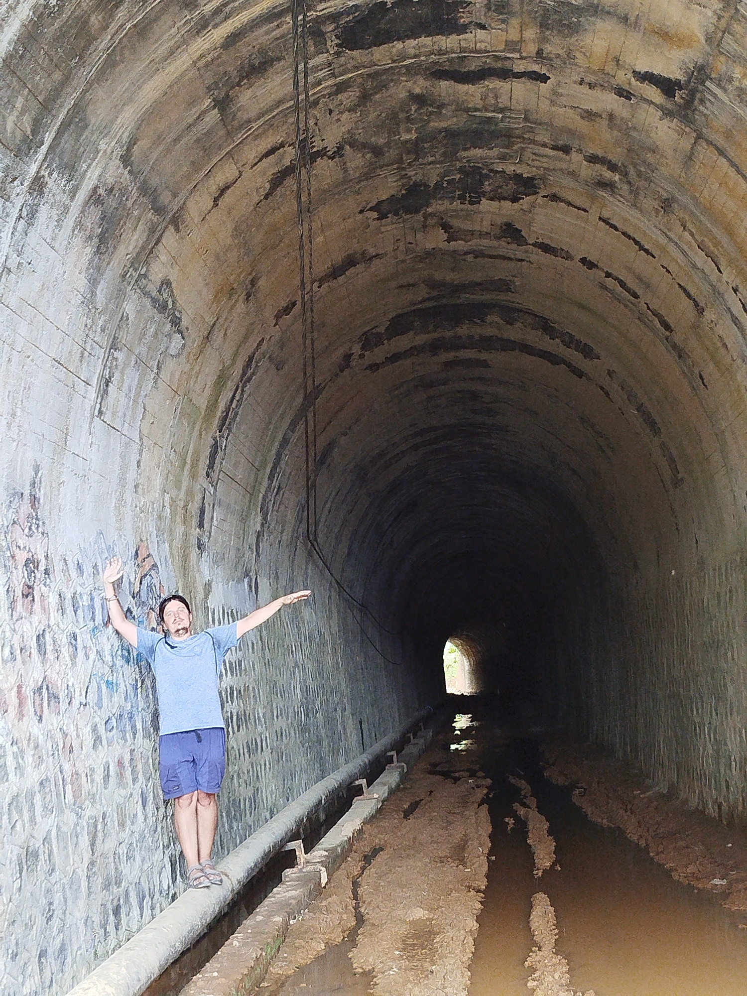 A person stands on a pipe within a tunnel. The tunnel has a brick or stone arched ceiling and walls. The tunnel floor is covered in mud and water. Light is visible at the end of the tunnel. There is a cable or rope hanging from the ceiling. The person is wearing a blue shirt, blue shorts, and sandals. They have their arms outstretched. The location is the city of Đa Thọ, Vietnam.