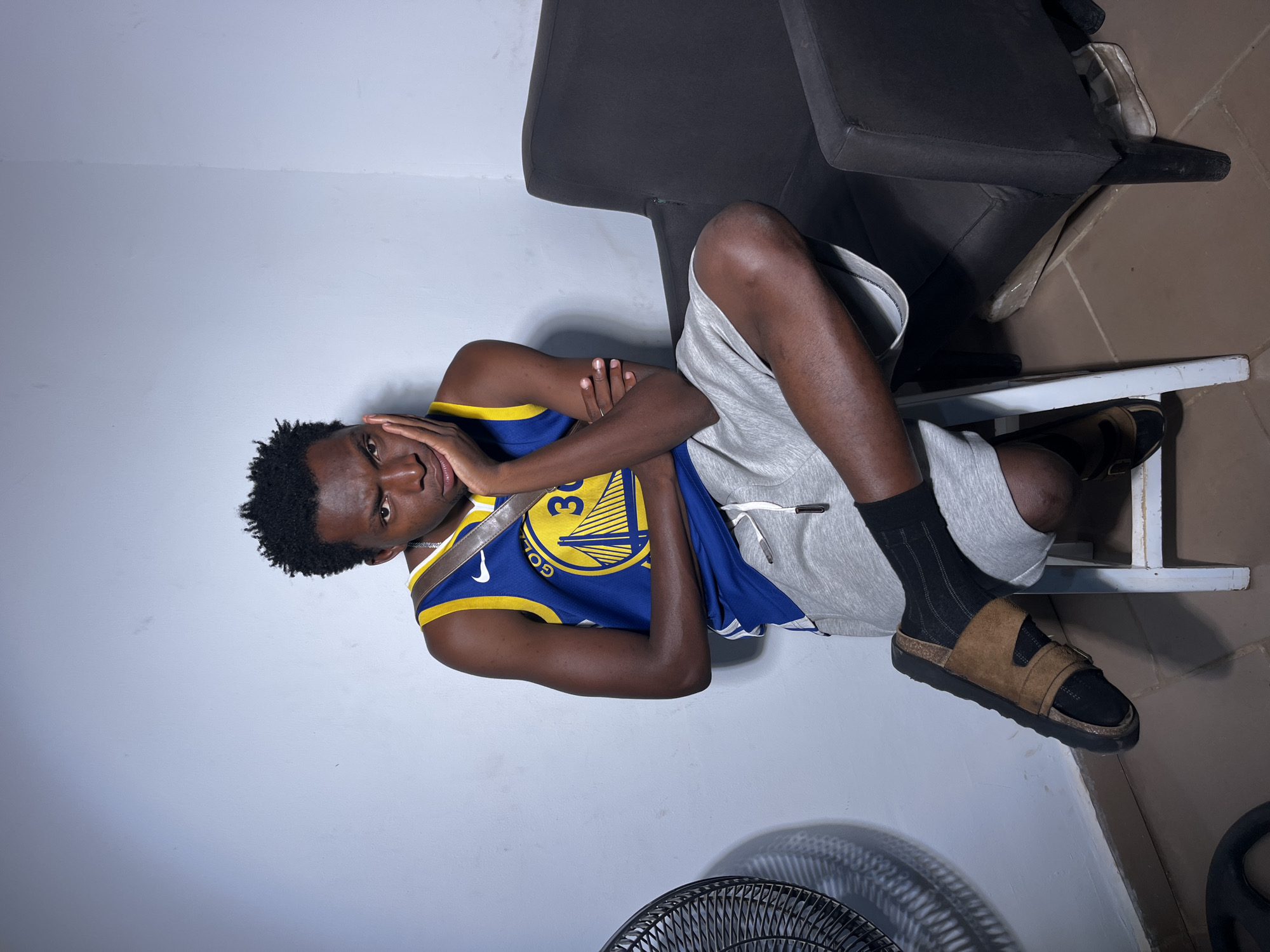 A young Black male sits on a white stool in an indoor setting, possibly a room with a light blue wall. He is wearing a blue and gold Golden State Warriors basketball jersey with the number 30 on it, gray athletic shorts, black socks, and brown sandals. He has his left arm resting on his knee and his right hand propped on his chin, with a pensive expression on his face. To his right, a dark gray armchair is visible. To his left, a black fan is partially in view. The flooring appears to be tiled in a reddish-brown hue.