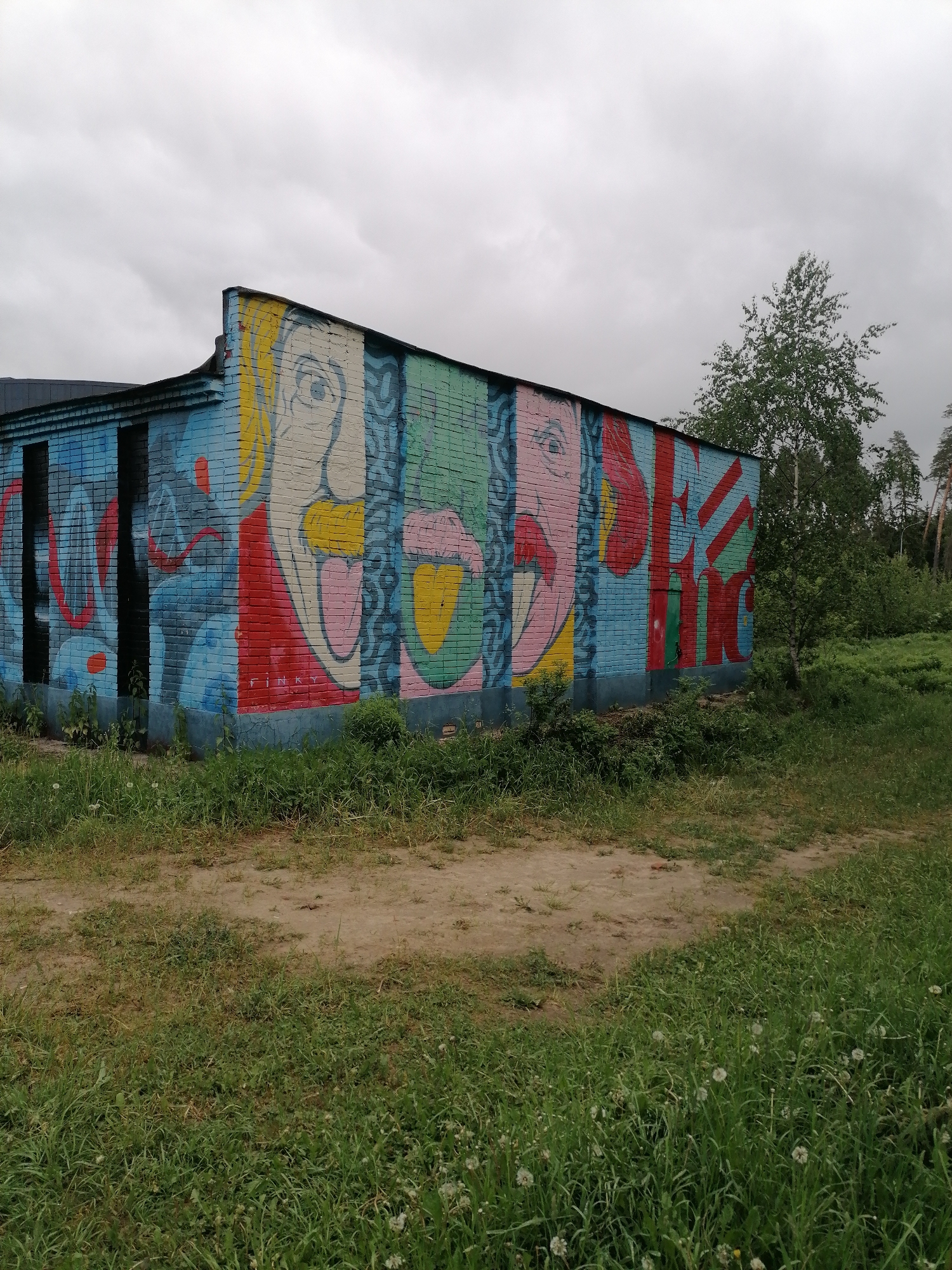 The image depicts a small, rectangular brick building located in an unkempt, grassy area under a cloudy sky in Unknown, Unknown. The building is adorned with colorful graffiti art, featuring stylized faces and lettering. Overgrown vegetation partially obscures the base of the structure. A dirt path runs in front of the building, and beyond it, a small tree and taller trees are visible in the background.