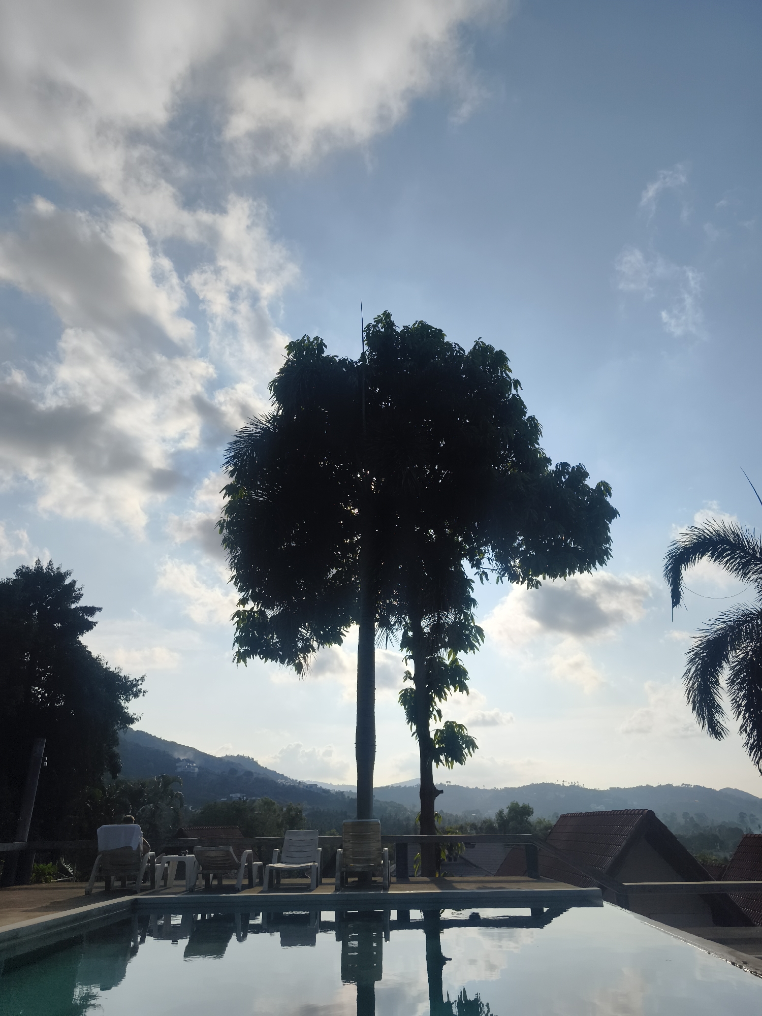 The image shows a view over a swimming pool at a location in บ้านบางรักษ์, Thailand. In the background are hills and rooftops. The sky is partly cloudy with blue patches. In the foreground are lounge chairs beside the pool, and trees are partially silhouetted against the sky. The water in the pool reflects the sky and the trees.