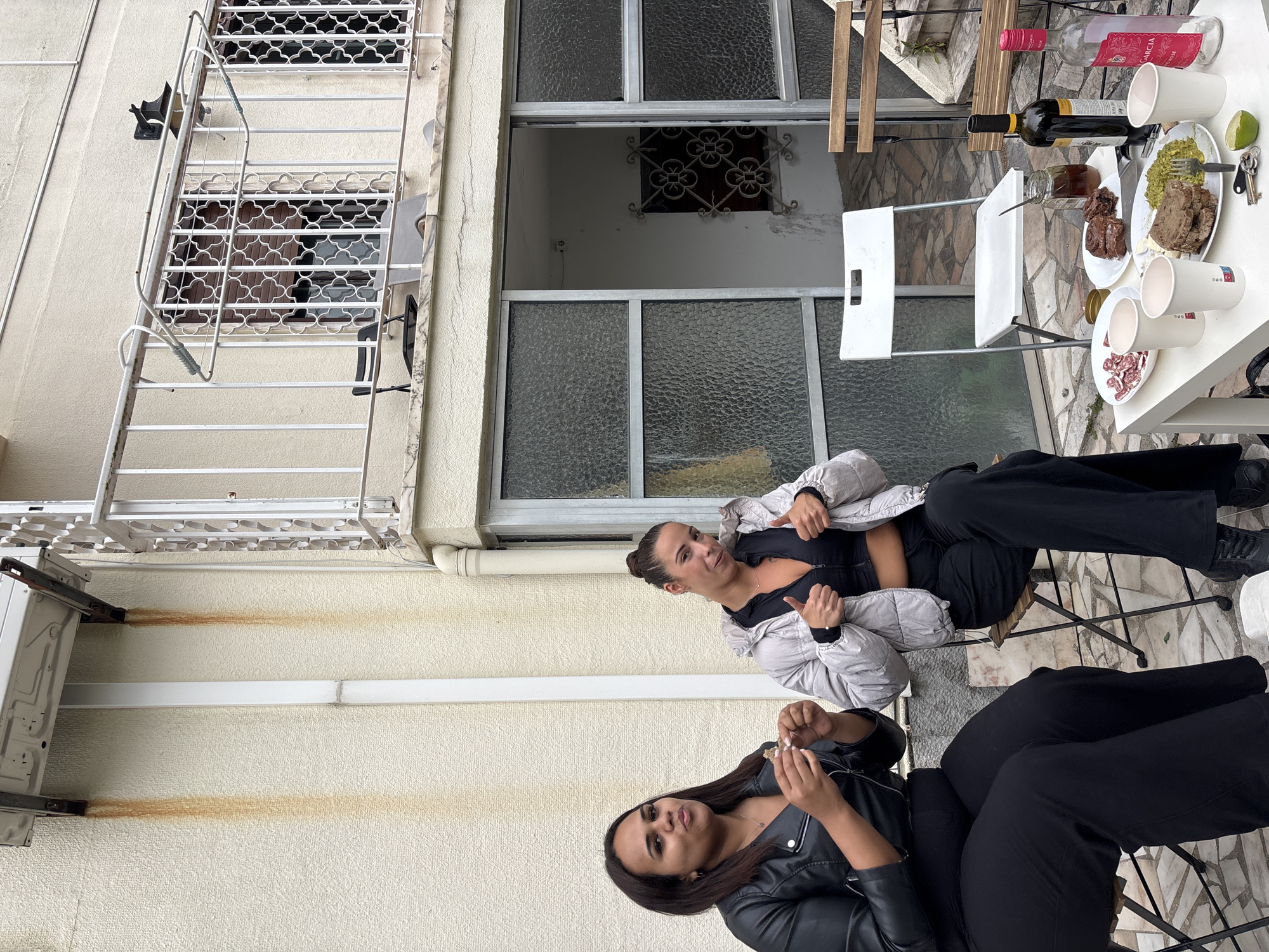 This image depicts an outdoor, casual gathering of two women on a balcony or patio in Lisbon, Portugal, under an overcast daytime sky.
The main subjects are two women. The woman in the foreground has long dark hair, wears a black leather jacket, and holds a small item in her hands, looking towards the camera with a relaxed expression. The woman seated opposite her, with dark hair tied back, is wearing a light grey quilted jacket over a black top and black trousers, giving a "thumbs up" gesture to the camera. Both appear at ease.
They are seated at a white table laden with food and drinks. Visible items include two bottles of wine: a red bottle with "CASTILLO DE PERELADA GARNACHA" printed on its label and a rosé bottle with "JOAN GARCIA" and "VINHO VERDE" text. Several white plastic cups, some with a small red logo, are scattered across the table. Plates hold various foods, including sliced cured meat (possibly salami), a portion of guacamole, and another dish with cooked meat. A lime wedge and a set of car keys are also on the table.
The setting reveals typical Portuguese architectural elements: light-colored textured building walls, windows with textured glass, and decorative wrought-iron security bars. A metal fire escape or ladder runs vertically along the left side of the frame. An outdoor air conditioning unit is mounted on the wall, with noticeable reddish-brown rust stains streaking down from its drainage points. The ground is paved with irregular, light-colored stone or marble tiles.