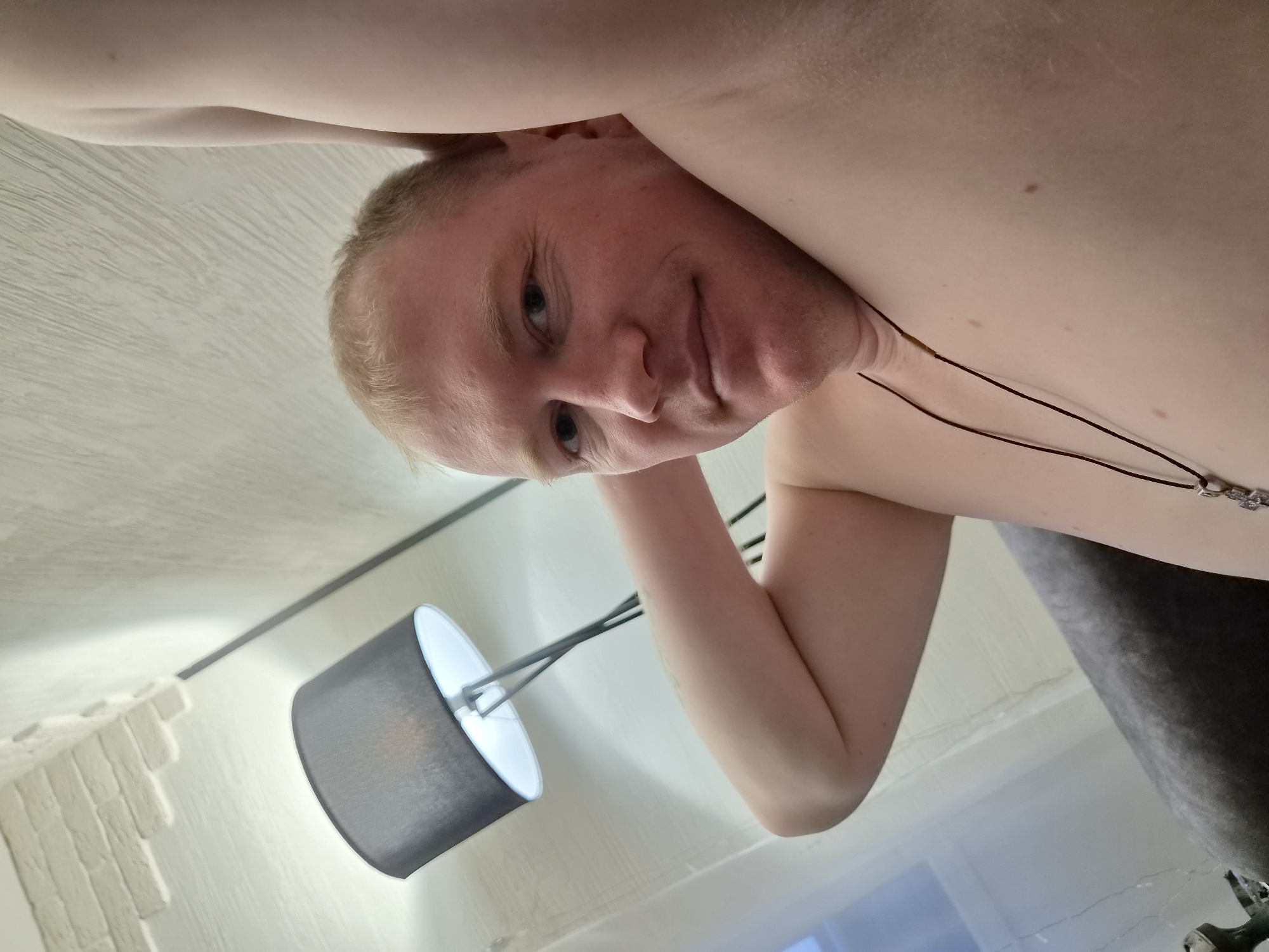 An inverted, low-angle indoor portrait features a shirtless, light-skinned man with short, light-colored hair and blue eyes. He looks directly at the camera with a subtle, friendly smile. He wears a dark cord necklace with a metallic, possibly cross-shaped, pendant. His left arm is visible, bent at the elbow.
The background, seen from a perspective looking upwards, shows a light-colored ceiling or upper wall on the left, part of which features a textured, possibly faux-brick, decorative surface. A modern floor lamp with a dark grey fabric shade and a visible white diffuser is positioned behind the man, casting light. To the right, a dark grey upholstered surface, likely a sofa or bed, is partially visible. The man appears to be leaning over or lying above the camera. The setting is an indoor space, possibly a living room or bedroom, in Chaikovsky, Russia. No text is visible in the image.