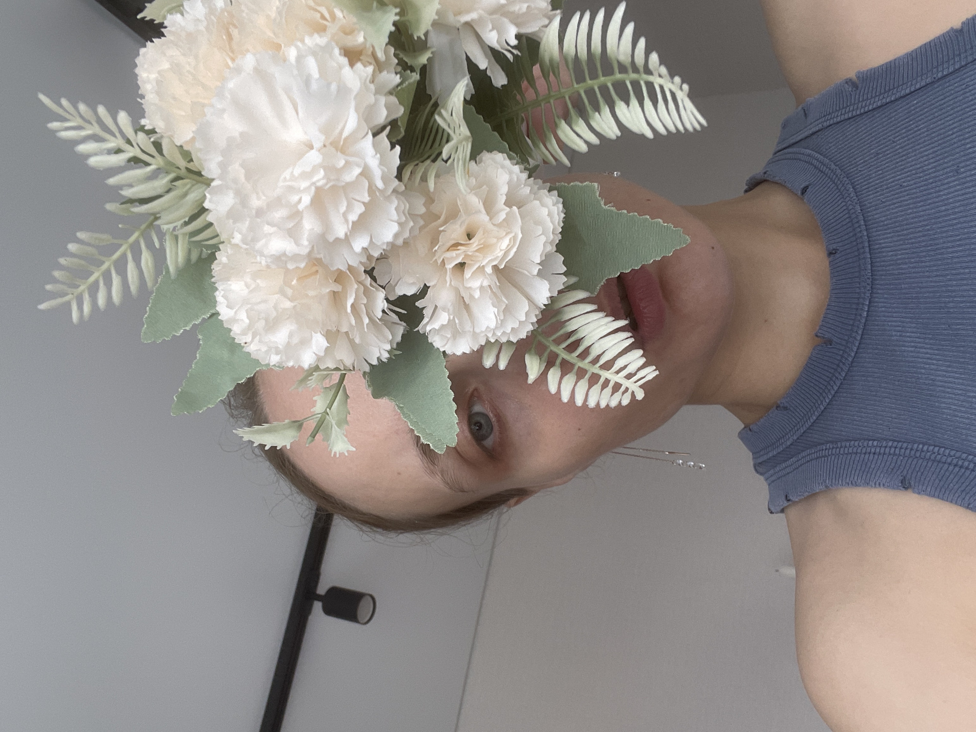 A head and shoulders shot shows a person with fair skin, wearing a blue ribbed tank top, holding a bouquet of pale, off-white flowers and green foliage. The person's face is visible, with one eye looking directly at the camera, and their mouth slightly open. A black adjustable lamp is visible on the left side of the image. The background is a light gray wall.