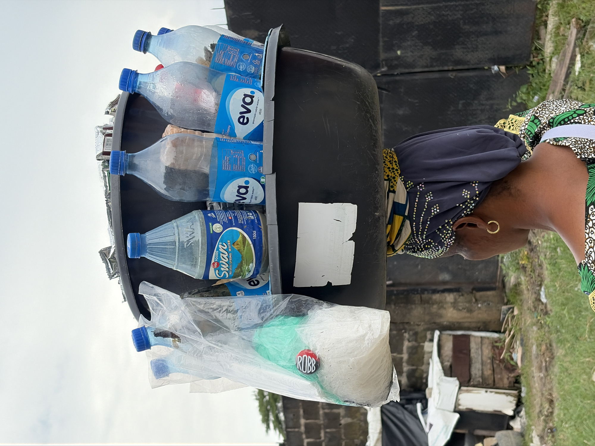 In Igboefon, Nigeria, a woman carries a large black basin filled with bottles of water and other items on her head. The basin contains at least five bottles of "Eva" premium table water, one bottle of "Swan" natural spring water, and other unidentified items in plastic bags. One bag contains a white powdery substance and a green object, with a round red and black object labeled "Robb" attached to it. The woman is wearing a headscarf and a colorful patterned top. The background shows a brick wall, wooden structures, and overgrown vegetation, suggesting an outdoor, possibly rural or semi-urban setting. The sky is overcast, indicating a cloudy day.
