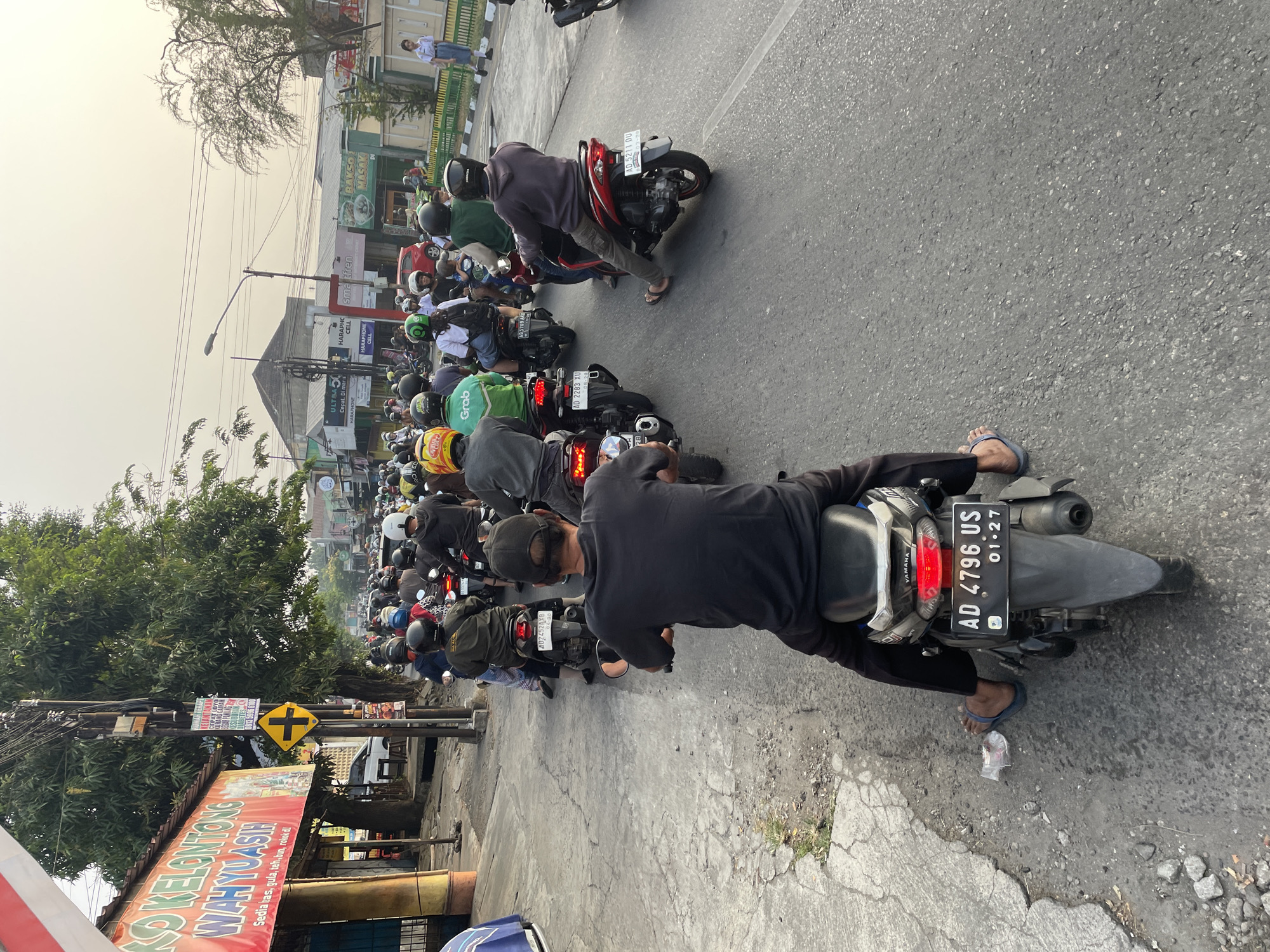 The image shows a busy street scene with a large number of motorcycles in traffic. The perspective is from behind a motorcyclist, looking forward at the procession. The road is paved and shows signs of wear and tear. On the left side of the road, there is a building with a red and white sign that reads "KO KELONTONG WAHYU ASIH". There are also trees and utility poles along the roadside. Several people are visible on motorcycles, most wearing helmets. The sky is overcast with a hazy, yellowish hue, suggesting either a sunset or air pollution. The overall impression is one of a congested urban environment.
