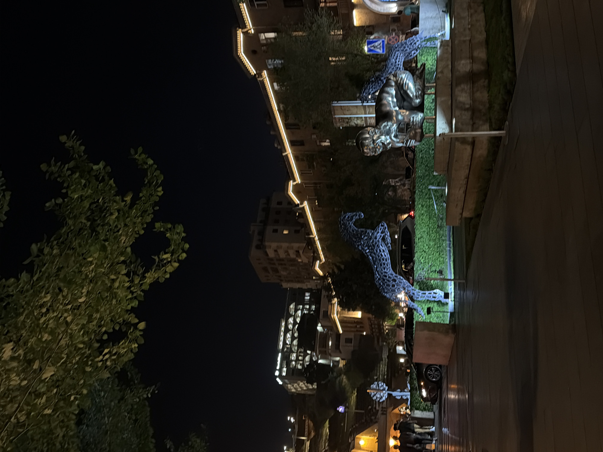 At night, a street scene in Yerevan, Armenia, features pedestrian activity alongside modern architecture, illuminated statues, and greenery. Buildings of varied heights are visible, some adorned with string lights along their edges. Statues of creatures with intricate details are centrally located within a green landscaped area, possibly a park. People are seen walking and standing near what appears to be a business or restaurant area. A road with vehicles runs adjacent to the pedestrian zone. The sky is dark, indicating a nighttime setting.
