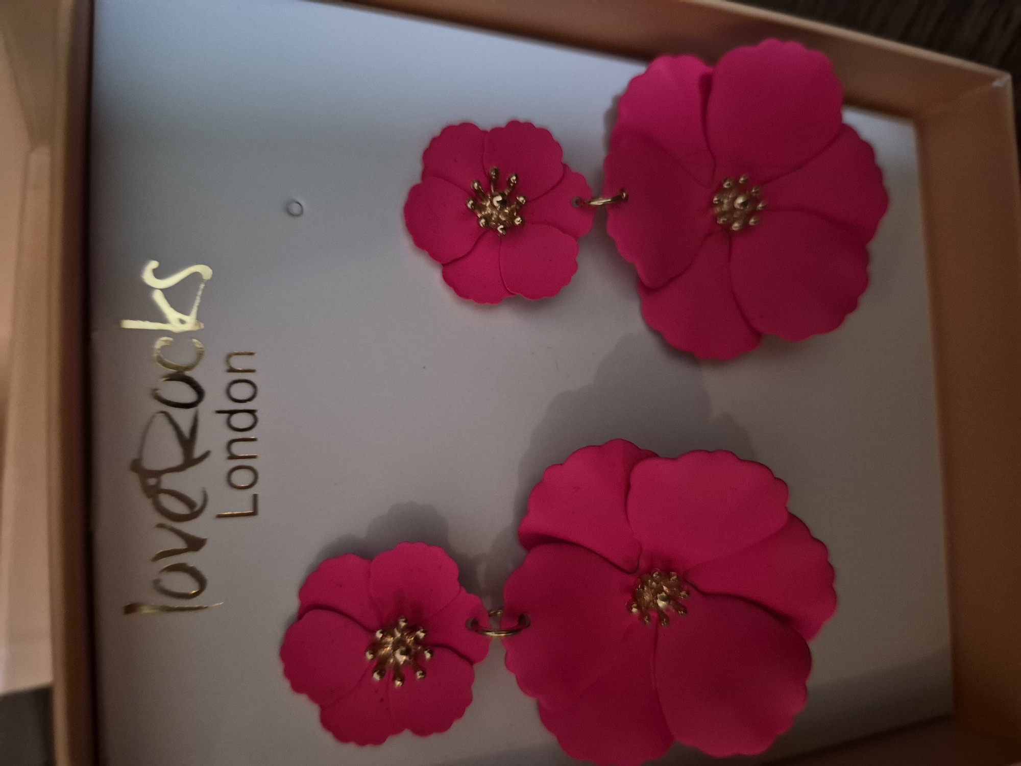 A pair of dangling earrings are displayed against a pale gray background, likely packaging for jewelry. Each earring features two bright pink, five-petaled flower shapes with gold centers. The flowers are layered, with a smaller bloom suspended above a larger one on each earring. The packaging is framed by a light brown border, and a gold logo reads "loveRocks London" at the top. The scene is indoors, with the lighting suggesting artificial illumination. There are no people present.