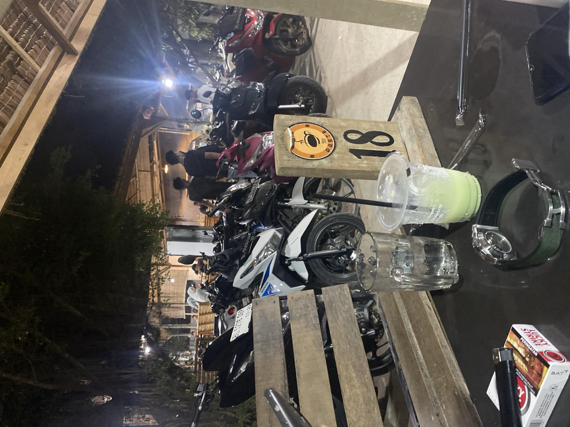 The image shows a table on the street in Gampongaceh, Indonesia, with a green smoothie in a plastic cup, a glass, a watch, a pack of cigarettes, and a lighter. Several motorcycles are parked near the table. People are sitting at tables in the background. It's nighttime.