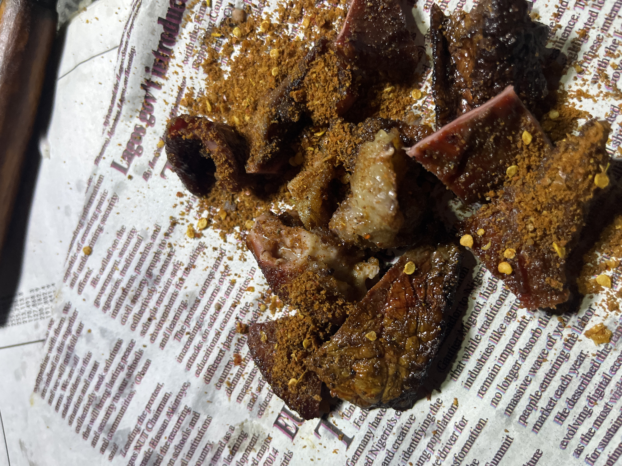 The image shows pieces of cooked meat, seasoned with a reddish-brown spice blend that includes chili flakes, scattered on a newspaper. The newspaper appears to be a Nigerian publication, with visible headlines and text related to the Lagos government and Nigeria. The meat is dark brown and appears to be grilled or roasted. The scene is likely a meal served on top of a newspaper, which is a common practice in some parts of Nigeria, including Abuja. There are no people visible in the image. The lighting suggests it was taken indoors, possibly in a domestic setting or a casual eatery. The overall impression is that of a simple, flavorful dish, likely enjoyed as a snack or part of a meal. The newspaper serves as a rustic serving platter, adding a cultural element to the presentation.